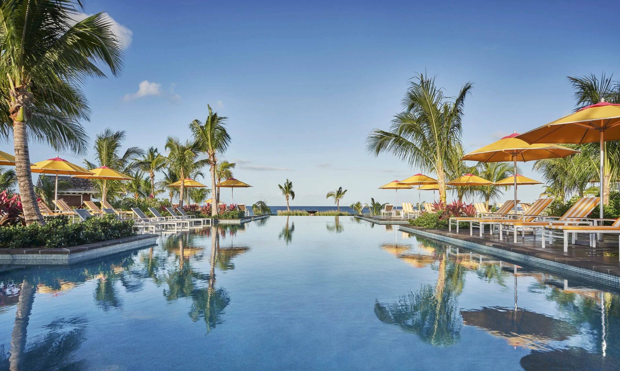 Swimming pool surrounded by yellow umbrellas and lounge chairs, palm trees, and a clear blue sky with clouds.