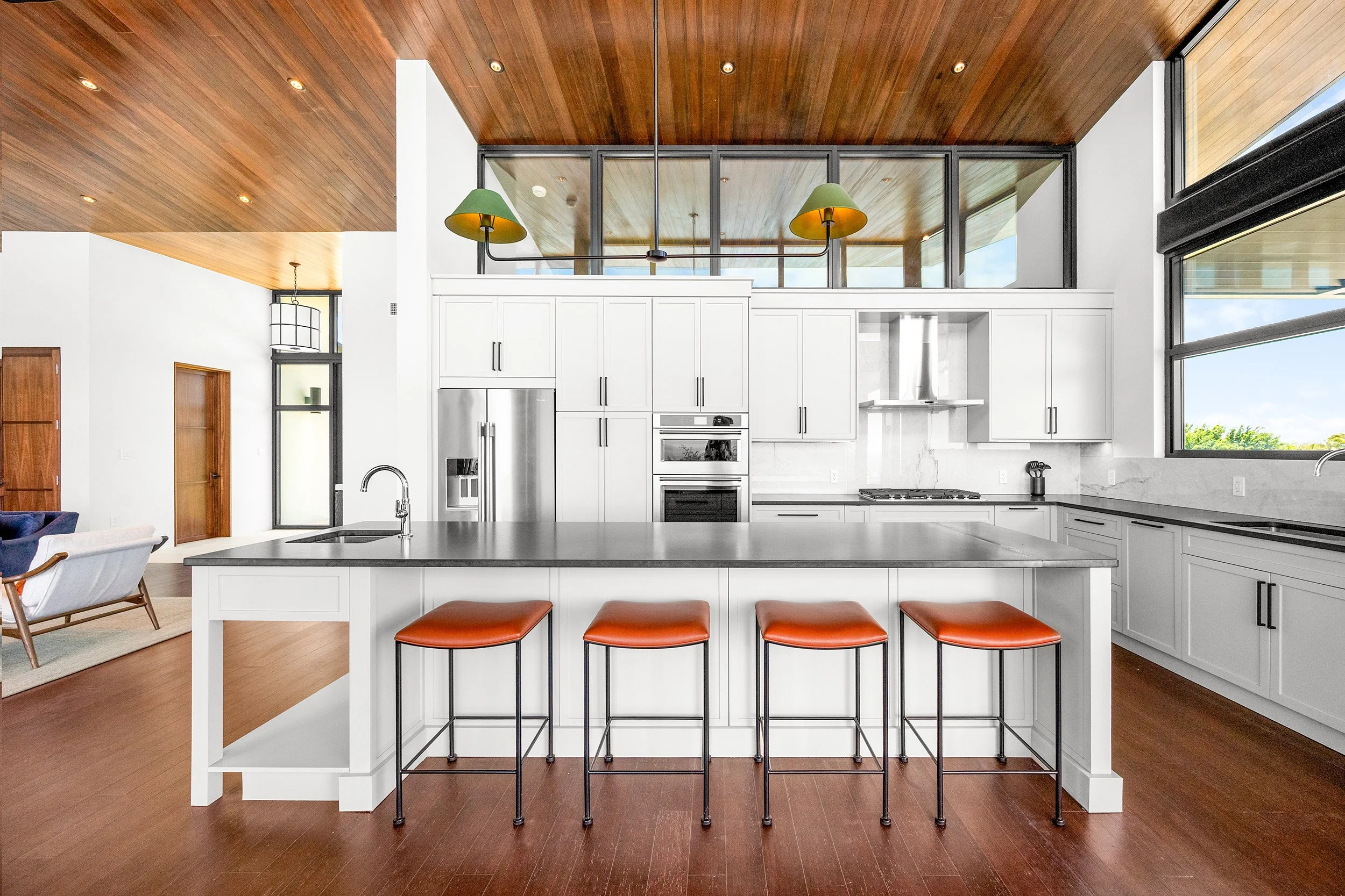 Modern kitchen with white cabinets, stainless steel appliances, large windows, and a wooden ceiling. Kitchen island with four bar stools and a view of the living room.