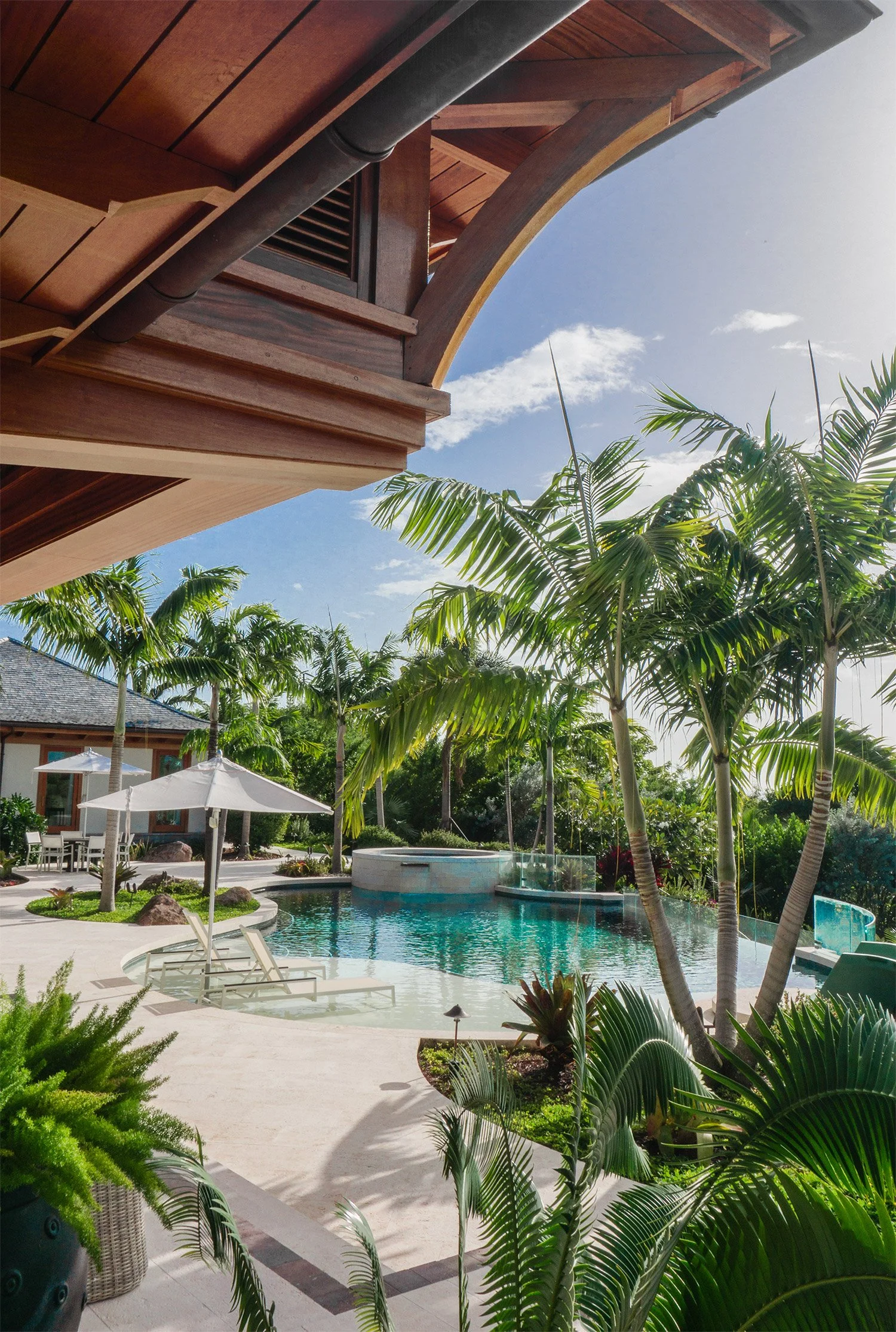 Pool area with lounge chairs, umbrellas, palm trees, and lush greenery under a bright sky