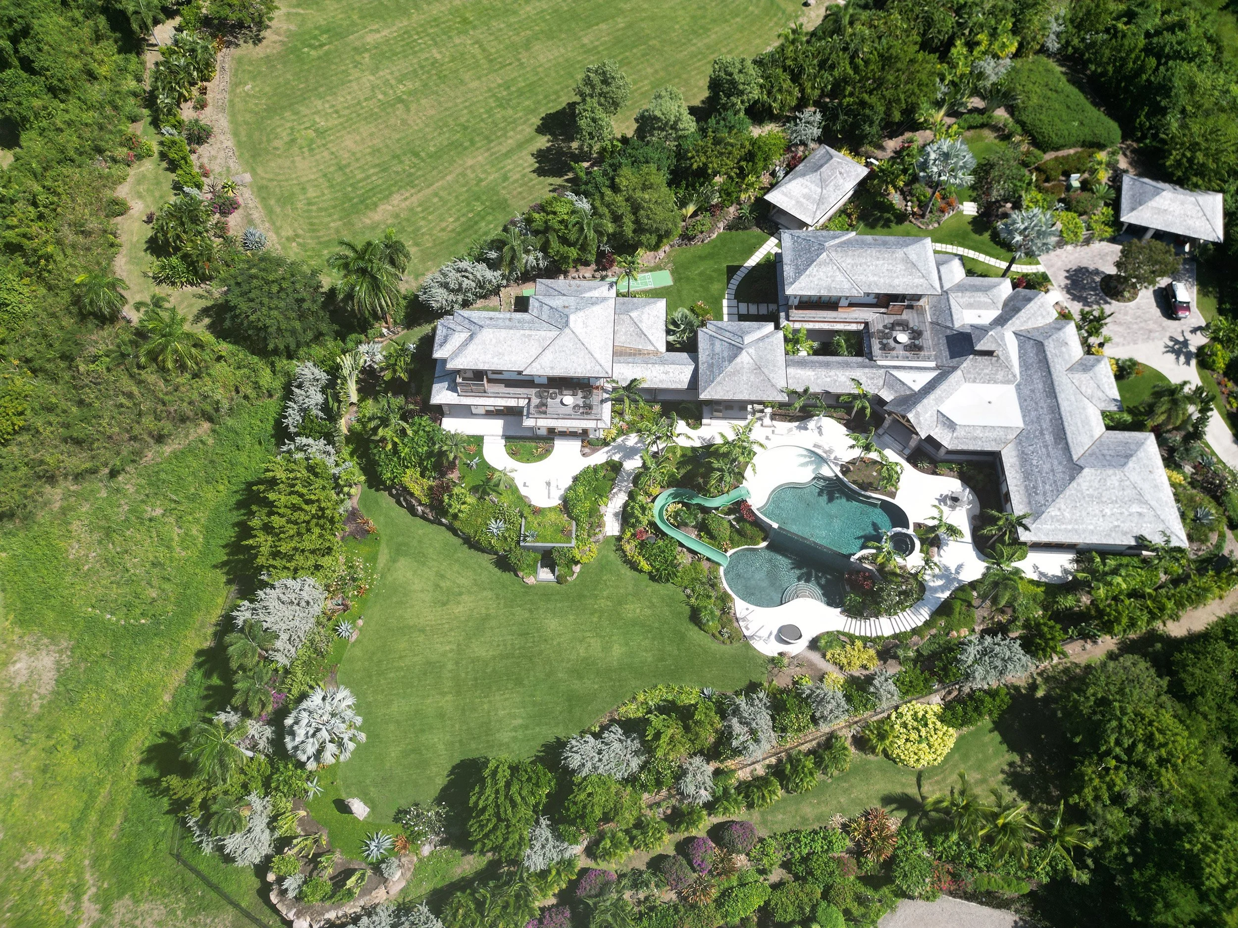 An aerial view of a large house with a swimming pool, surrounded by lush green trees and gardens, with a spacious lawn area nearby.