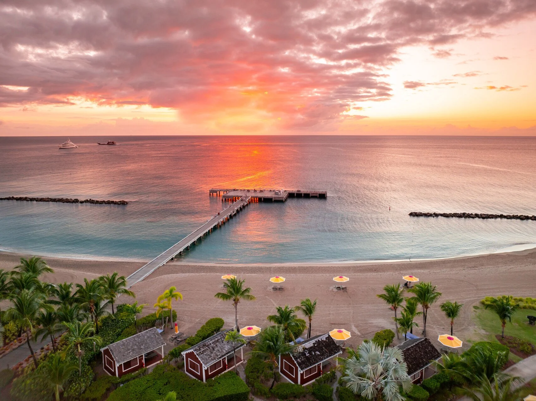A beach at sunset with calm water, a wooden pier extending into the ocean, boats in the distance, green palm trees, and beach umbrellas with chairs along the shoreline.