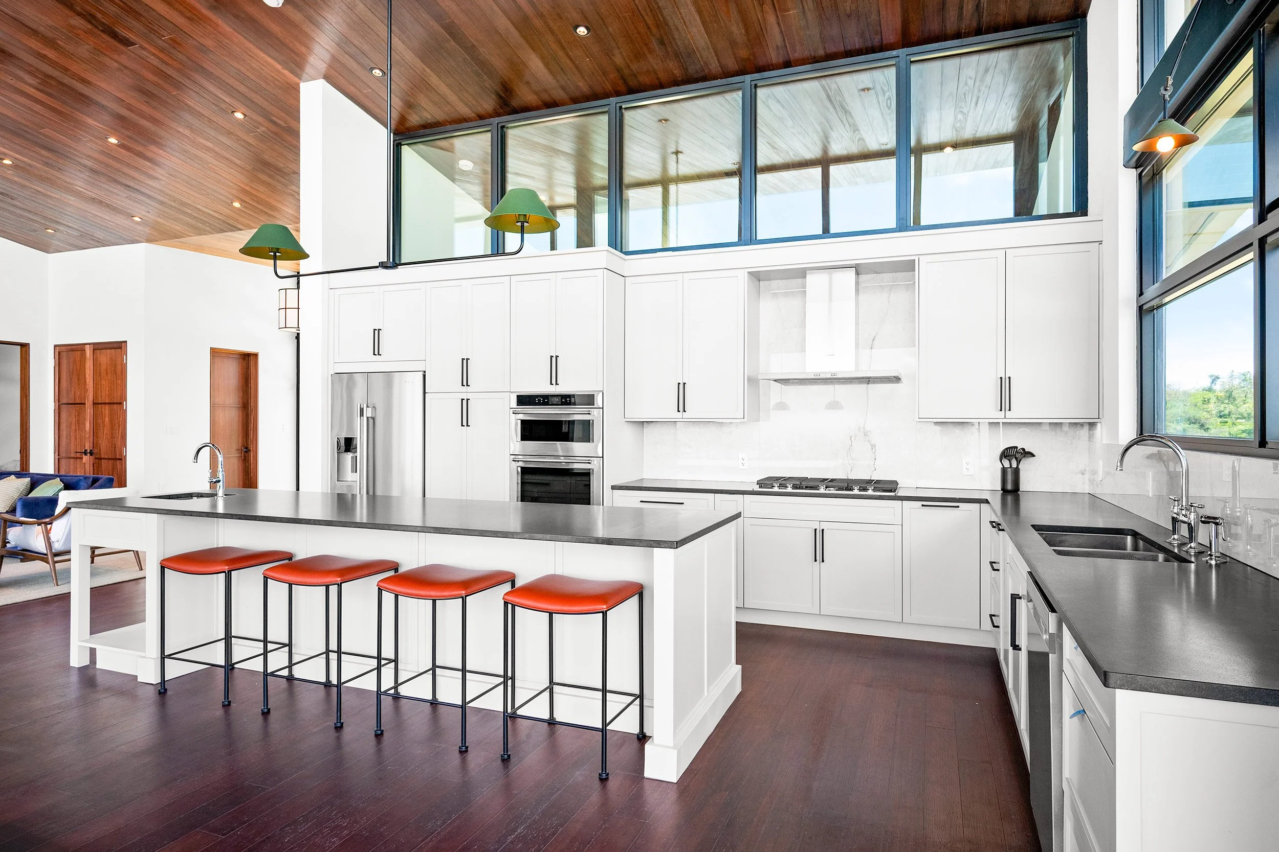 Modern kitchen with white cabinets, marble backsplash, stainless steel appliances, dark wood flooring, and a large island with four red barstools. Large windows and a high wooden ceiling with recessed lighting.