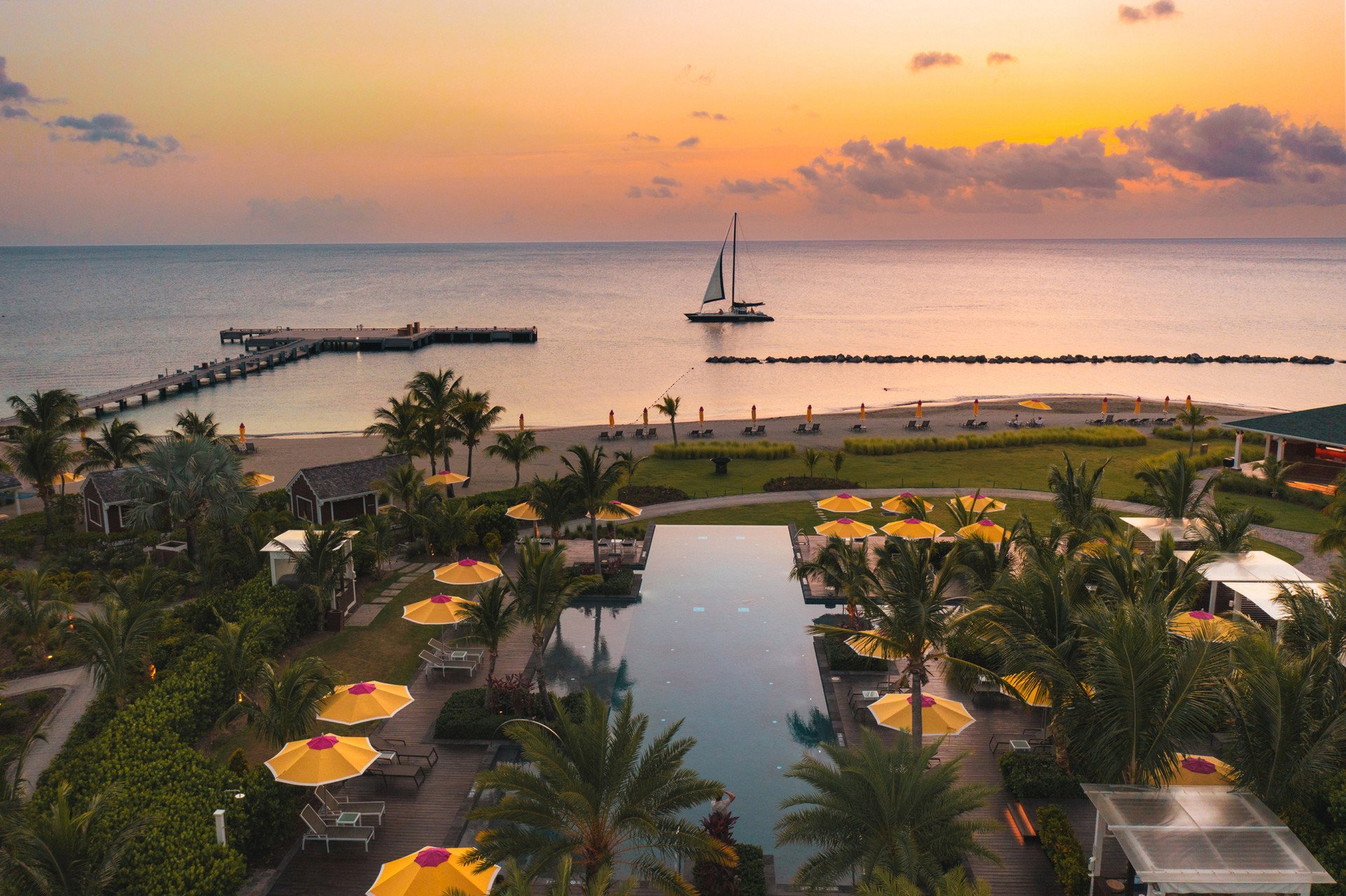 Sunset over a calm ocean with a sailboat near the horizon, a pier extending into the water, and a resort pool surrounded by yellow umbrellas and tropical palm trees.