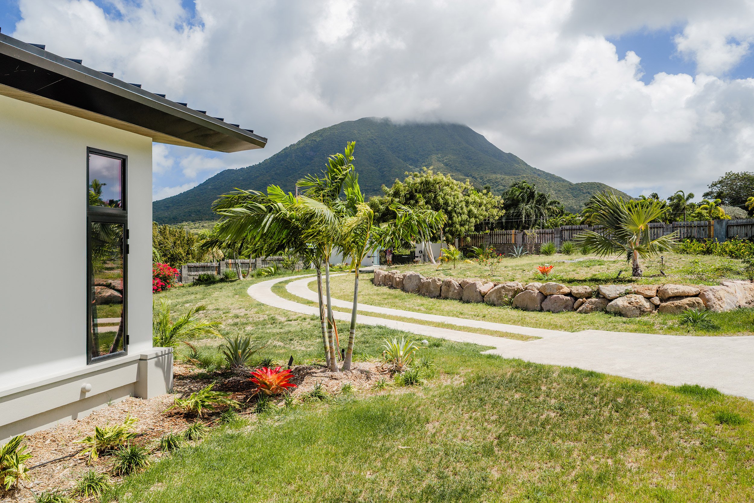 A modern house with a glass door and window, surrounded by a well-maintained garden with small trees, bushes, and colorful flowers, with a mountain in the background under a cloudy sky.
