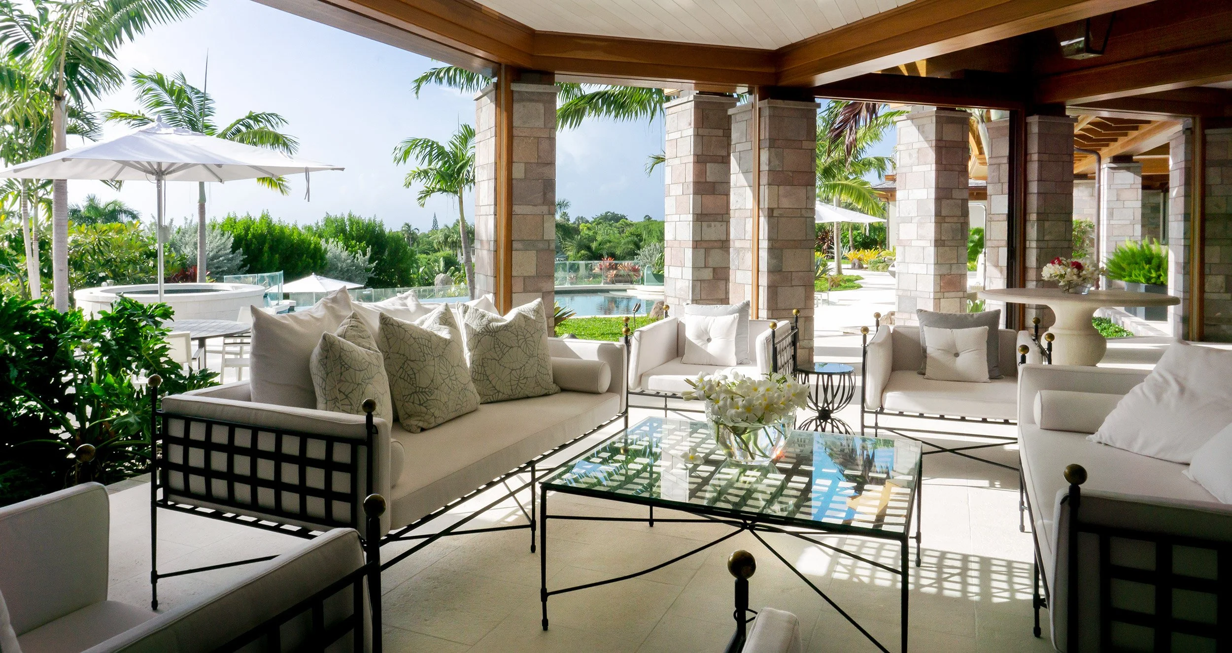 Indoor patio with white furniture, throw pillows, glass coffee table, surrounded by lush greenery and palm trees, with a view of a pool outside under a sunny sky.