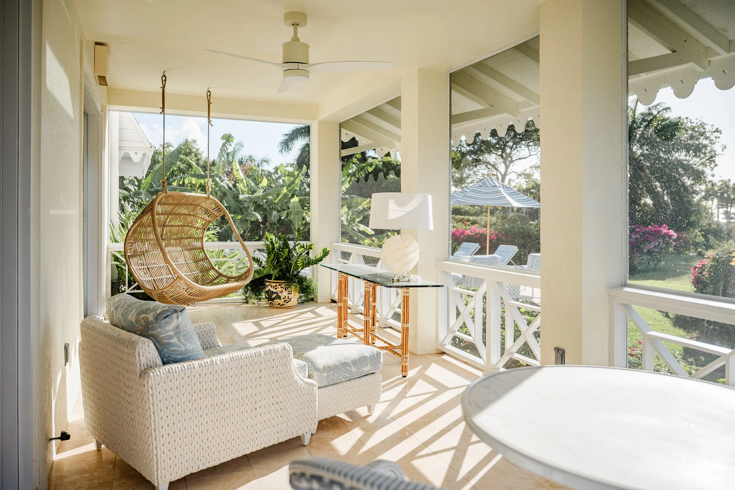 Sunlit screened porch with wicker hanging chair, white wicker armchair with blue patterned pillow, plants, glass table with white shell lamp, and views of lush greenery and a patio with lounge chairs and an umbrella outside.