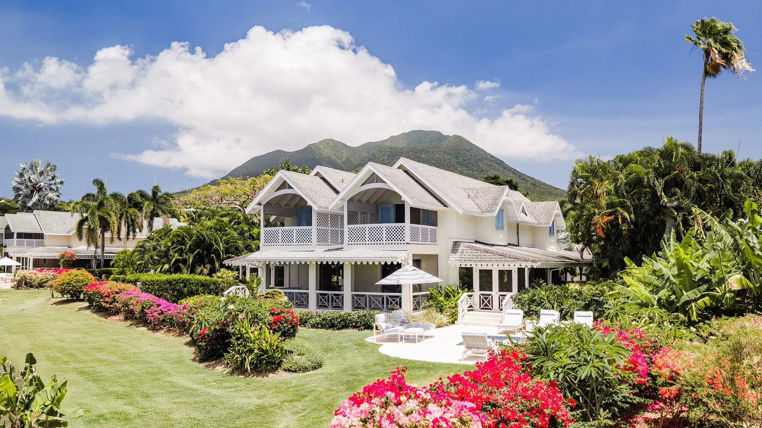 A bright, white, two-story house with a patio surrounded by lush green gardens, colorful flowers, tall palm trees, and mountains in the background under a partly cloudy sky.