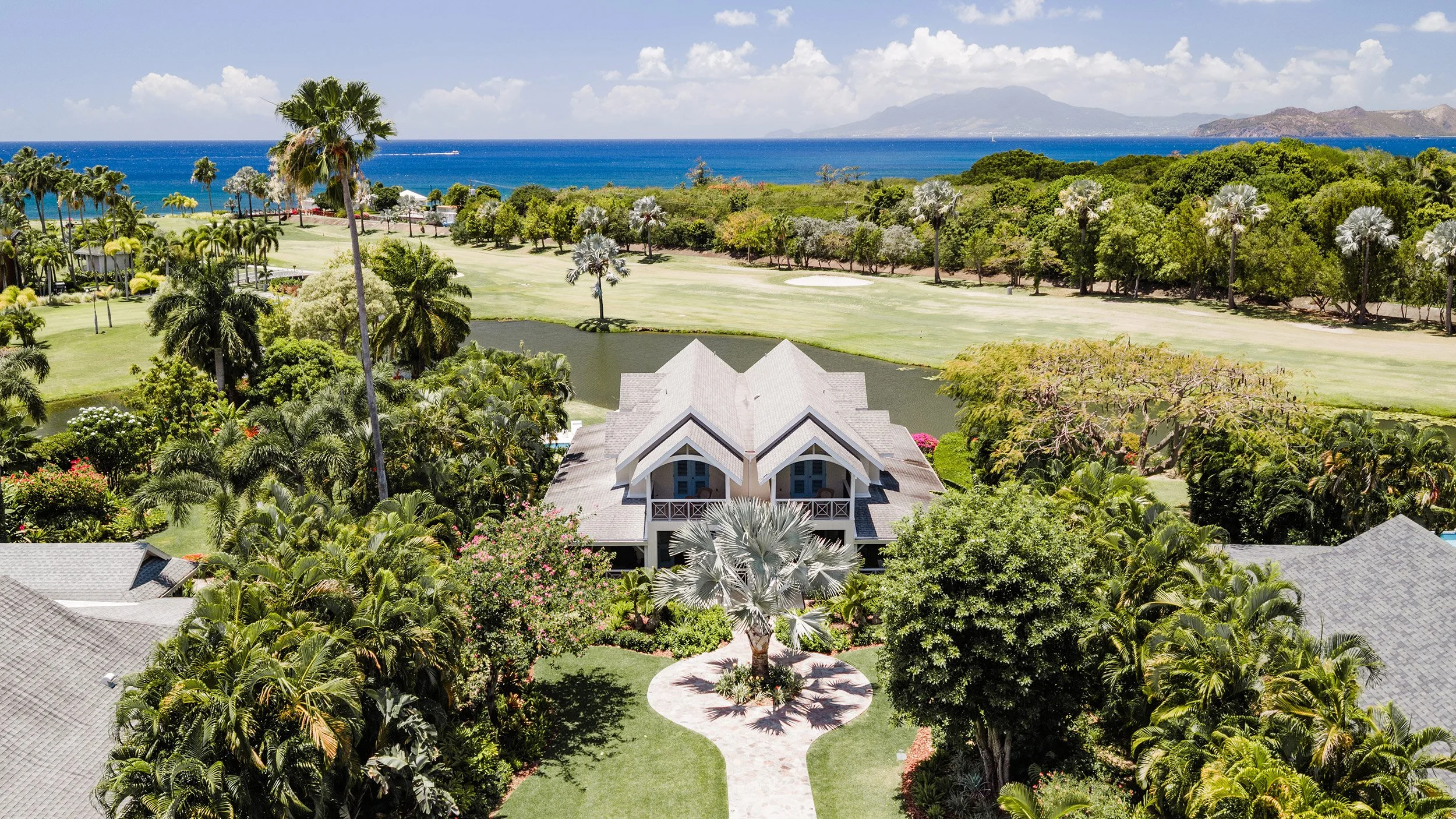 Aerial view of a coastal property with lush tropical landscaping, a golf course, a pond, and a house with a gray roof, overlooking the ocean and distant mountains.