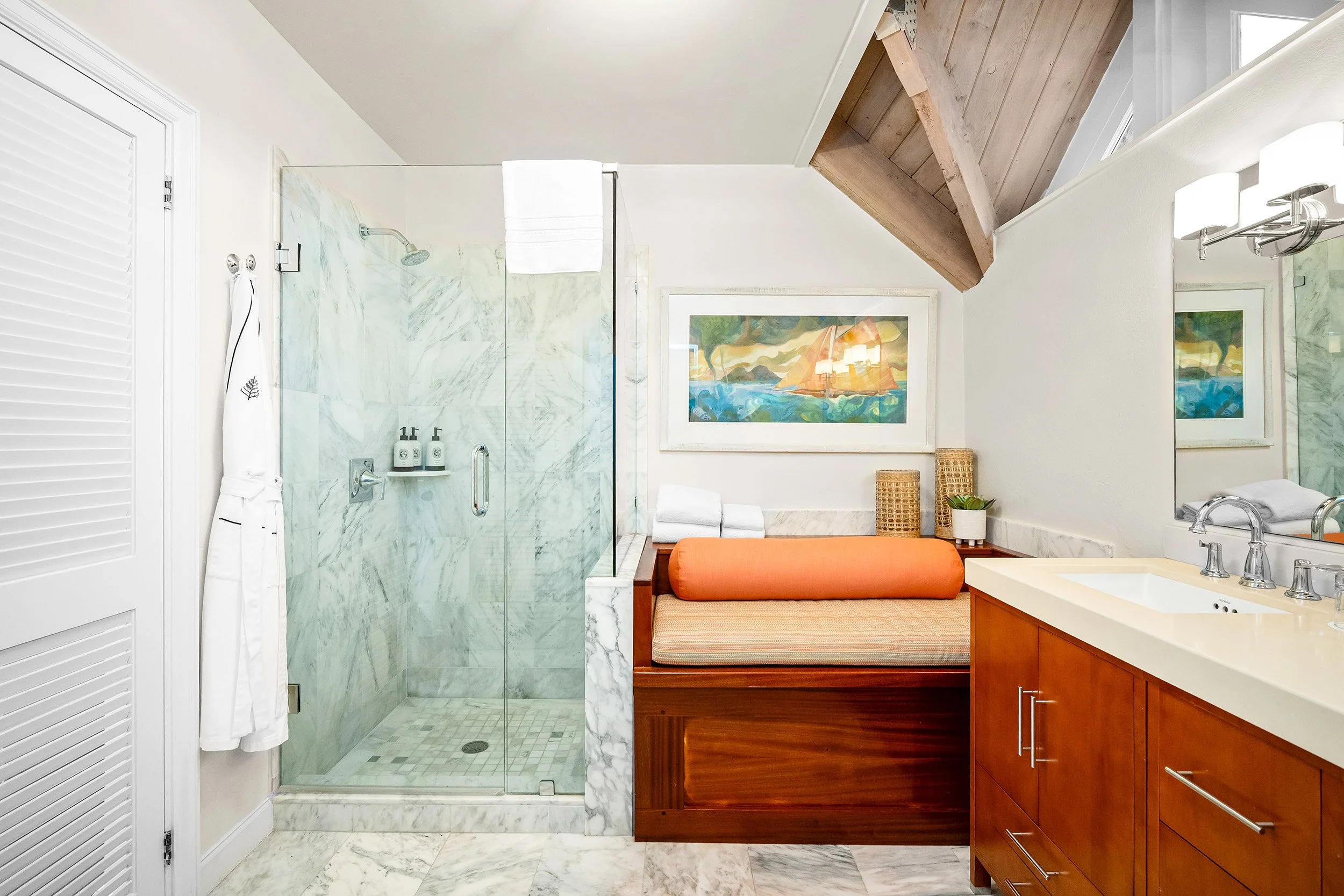 Bathroom with a walk-in shower with marble walls, a wooden bench with an orange cushion, a framed painting of a sailboat on the wall, and a vanity with a white sink and chrome fixtures.