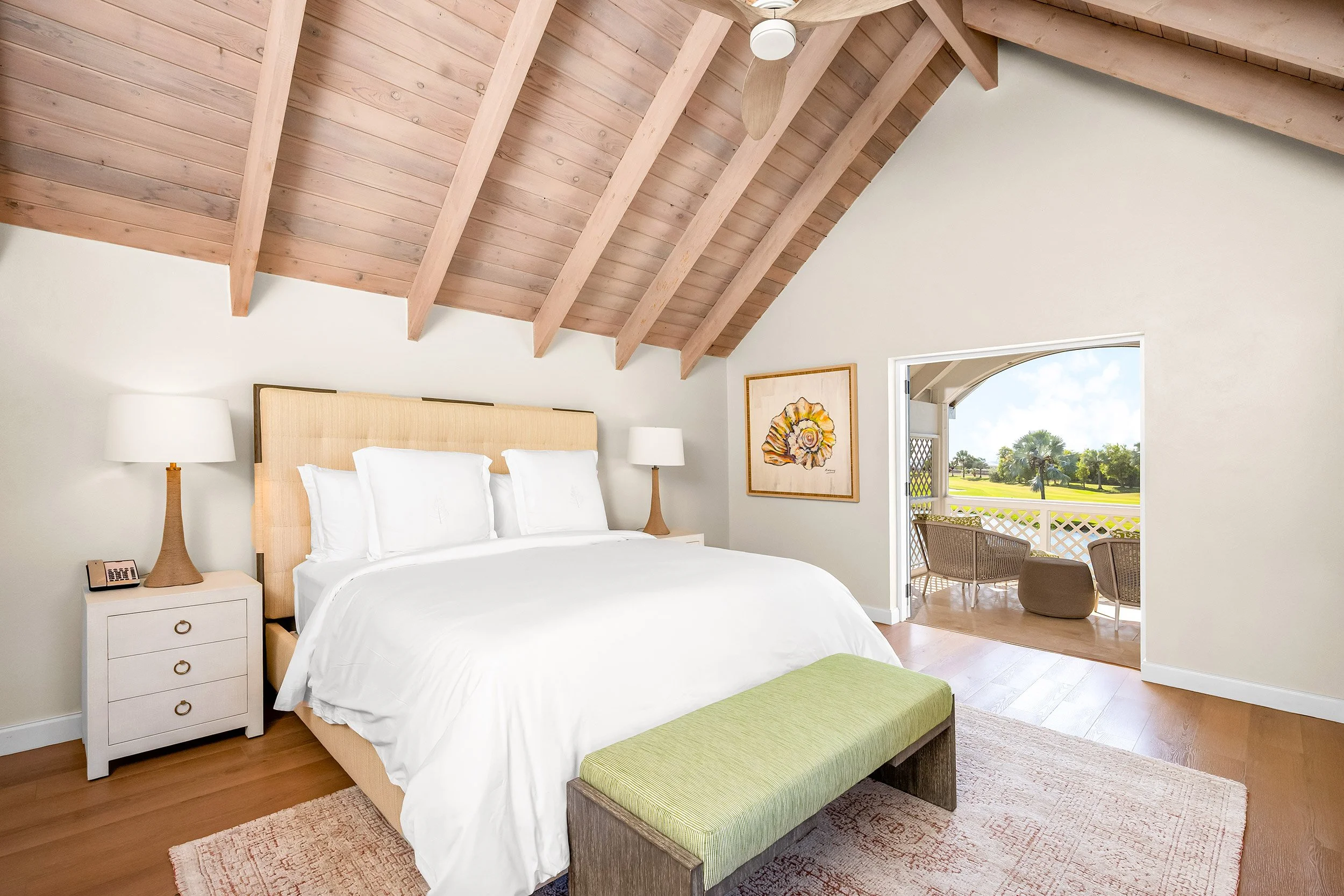 A cozy bedroom with a sloped wooden ceiling, a large bed with white linens and a beige headboard, matching bedside tables with lamps, a piece of artwork on the wall, and a small balcony with outdoor seating and a view of trees and a blue sky.