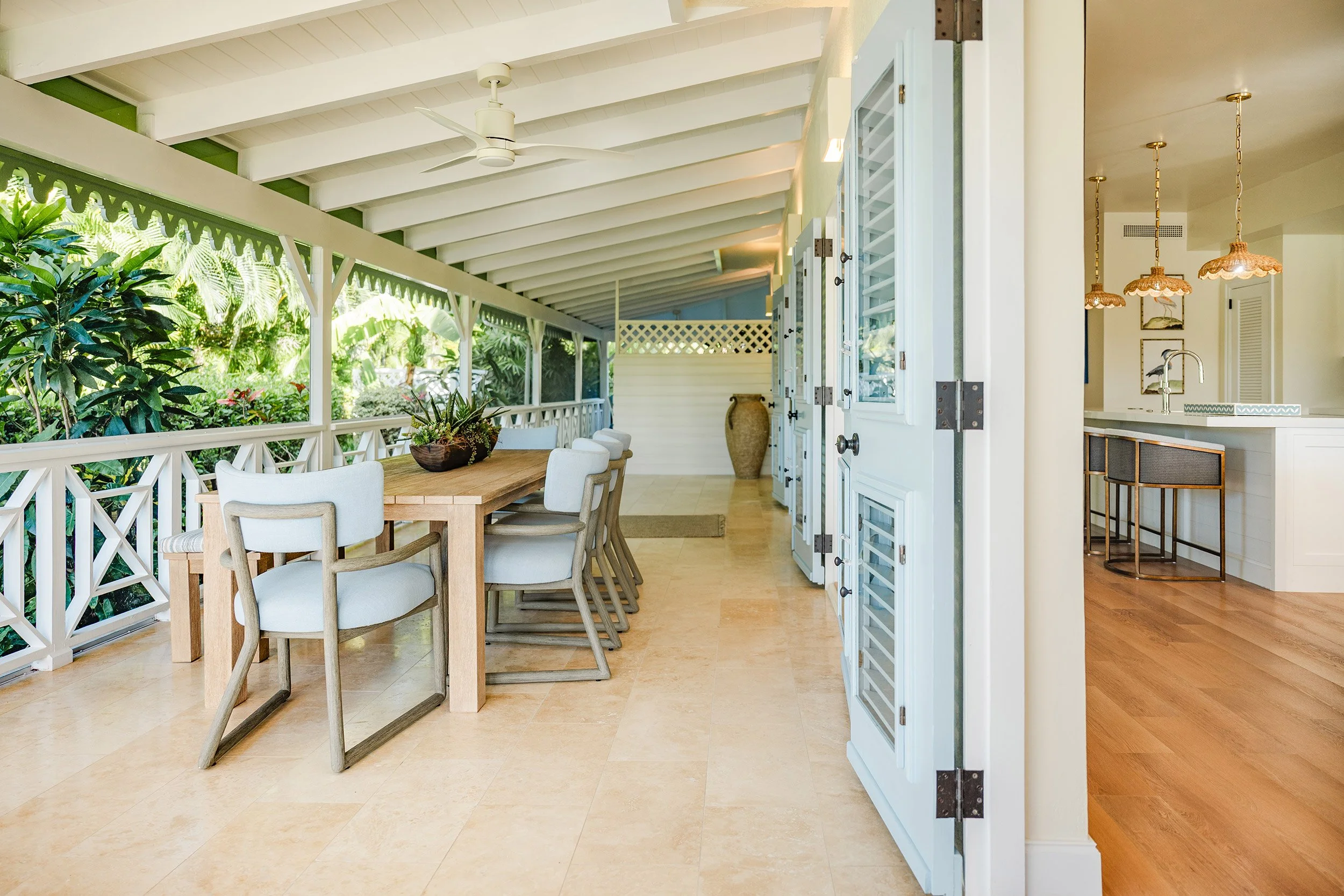 A covered outdoor patio with a dining table, chairs, and lush greenery view, adjacent to a modern kitchen with hanging pendant lights.