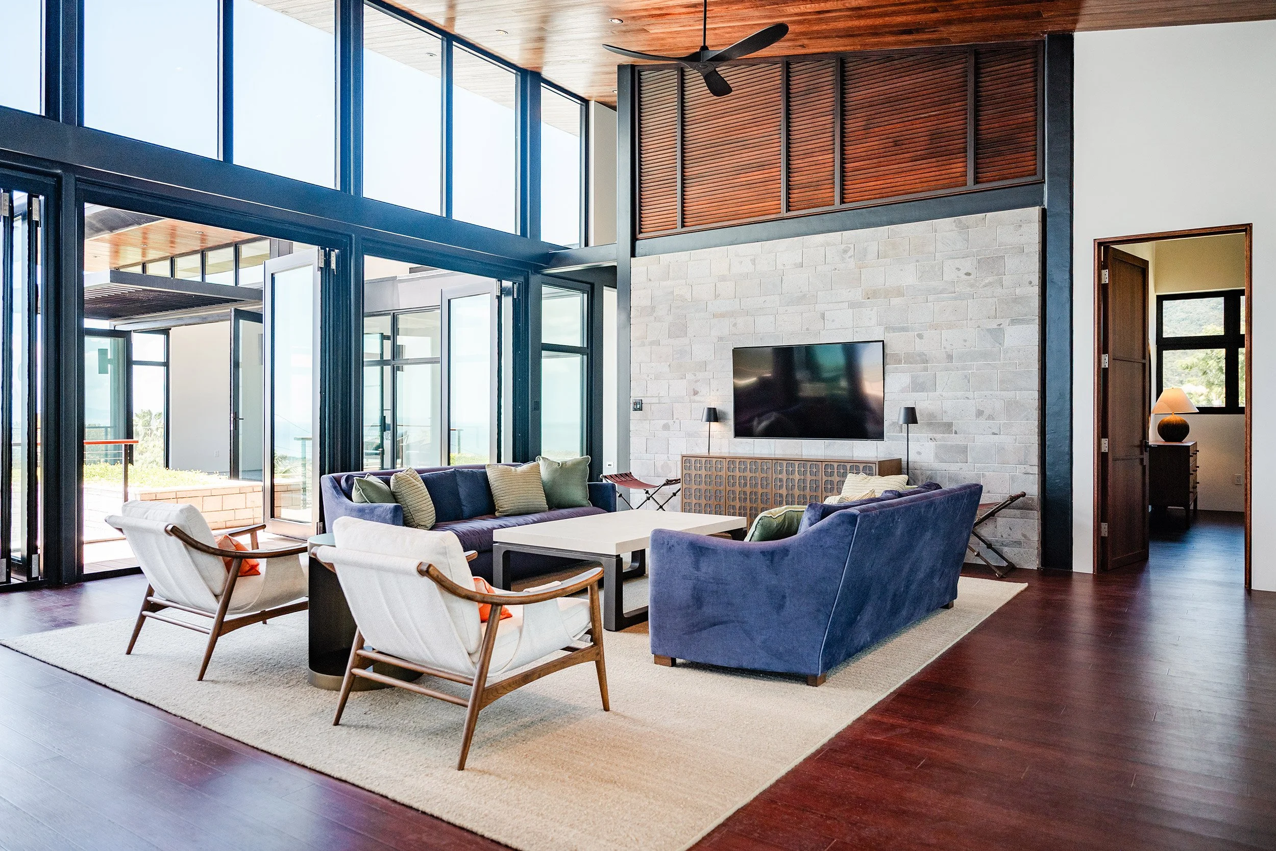 Modern living room with large glass windows, a stone fireplace, a wall-mounted TV, and a mix of contemporary seating including a sofa and armchairs, with wooden flooring and ceiling.