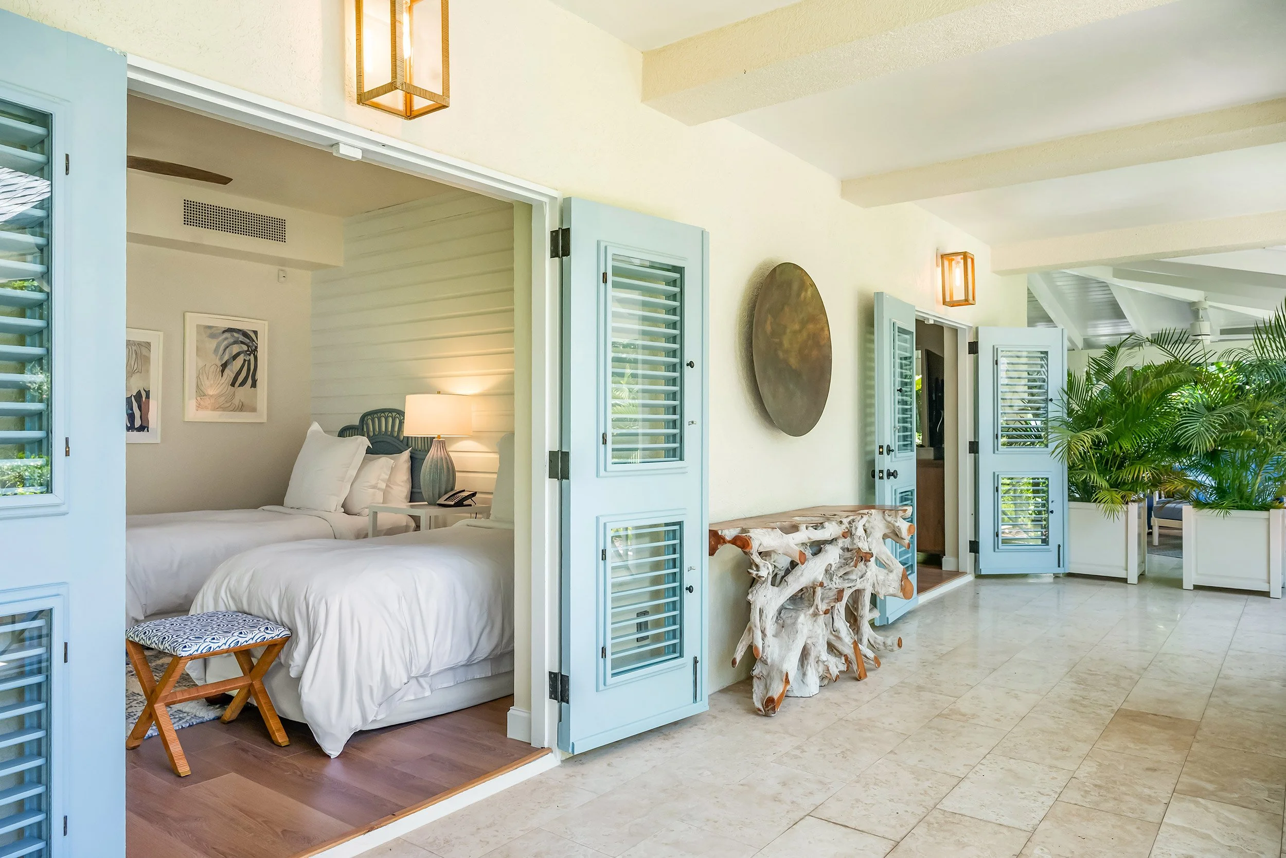 View of a bedroom with two beds, a nightstand with a lamp, and decorative artwork, separated from a living area with light blue shutters and a rustic wooden table, by blue shutters and door.