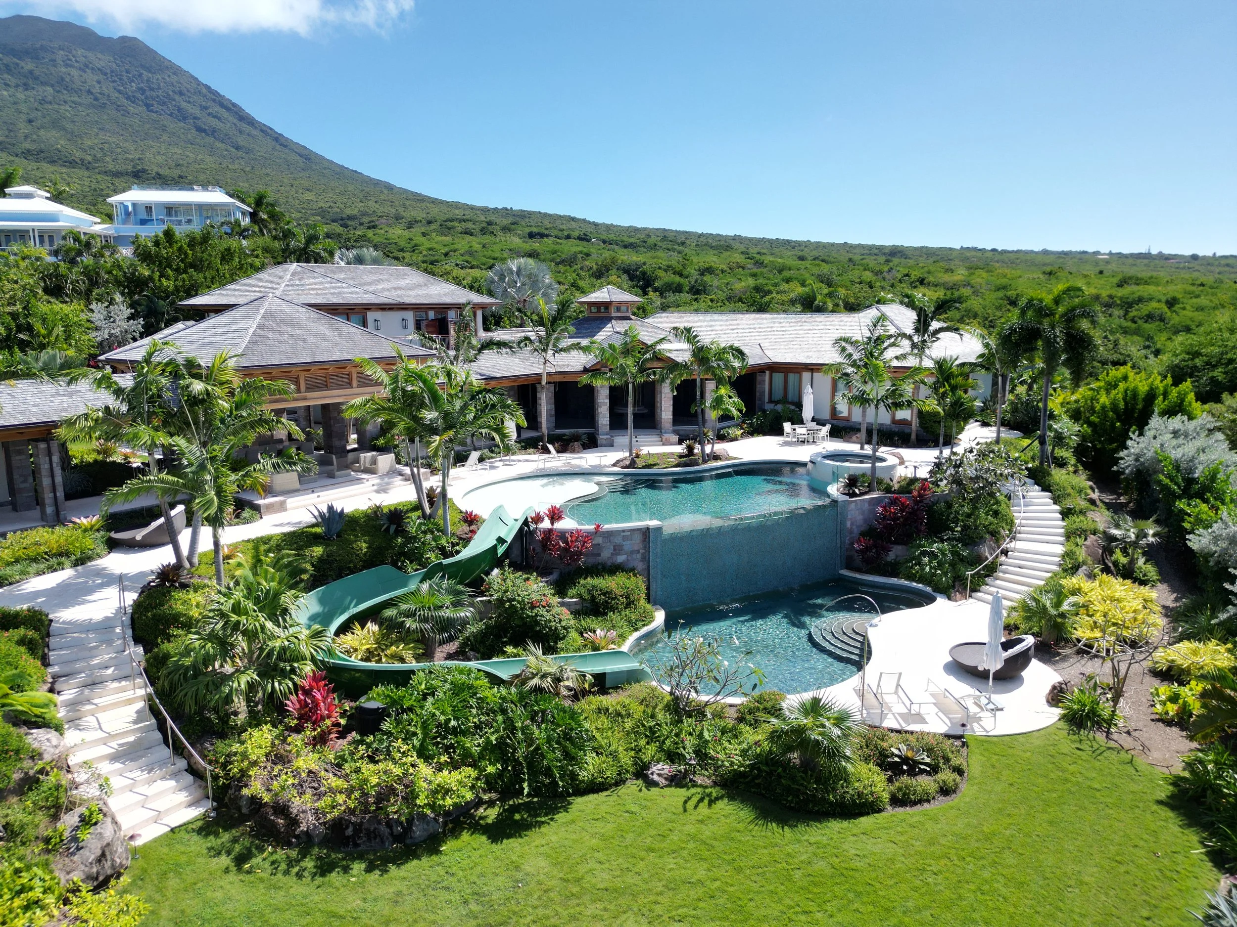Luxury house with a swimming pool, surrounded by lush greenery and palm trees, in a mountainous area under a clear blue sky.