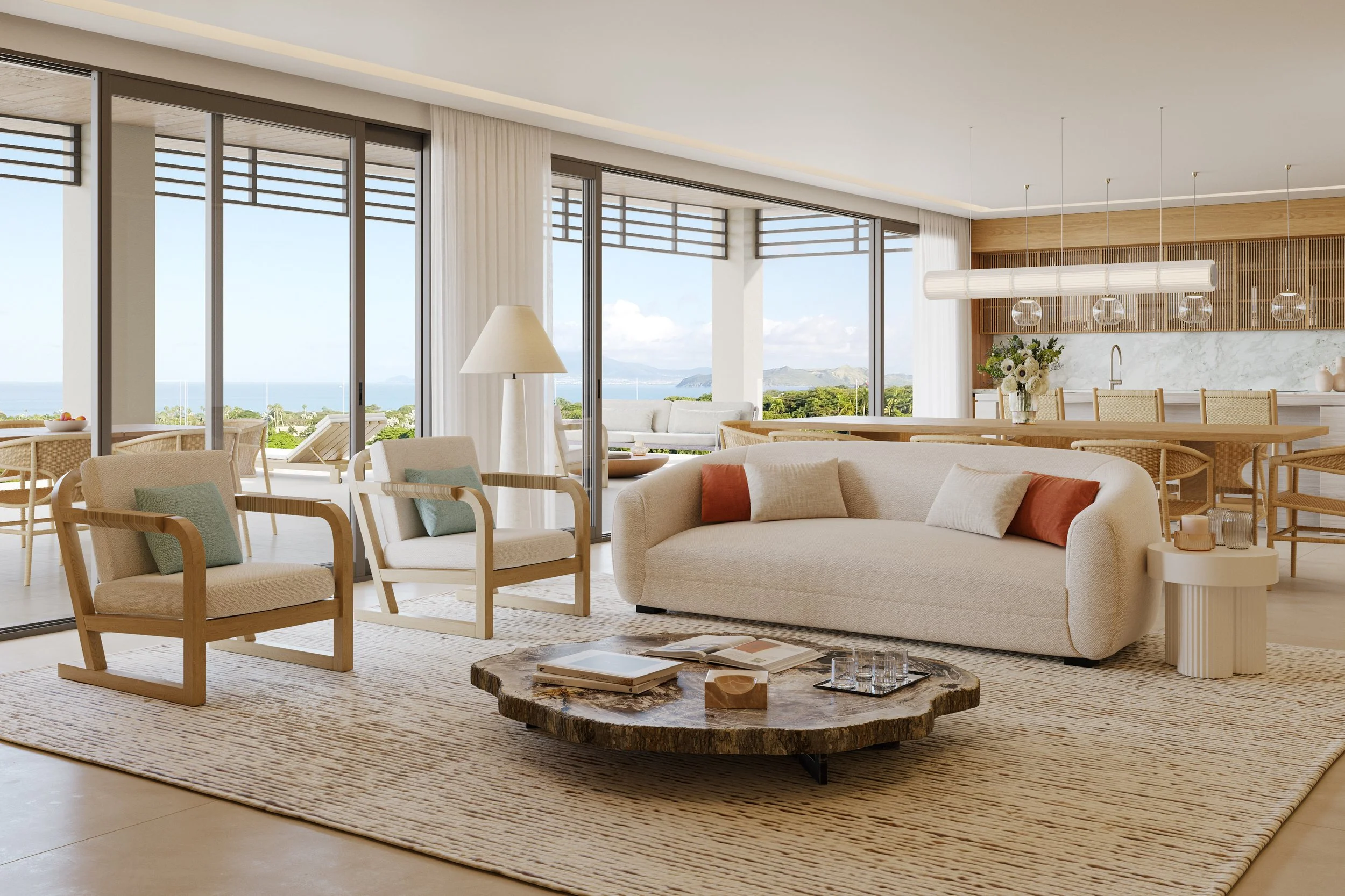Bright living room with large windows showing outdoors, neutral-colored sofas, a round coffee table with books and decorative items, and a view of a coastline with blue sky and mountains in the background.