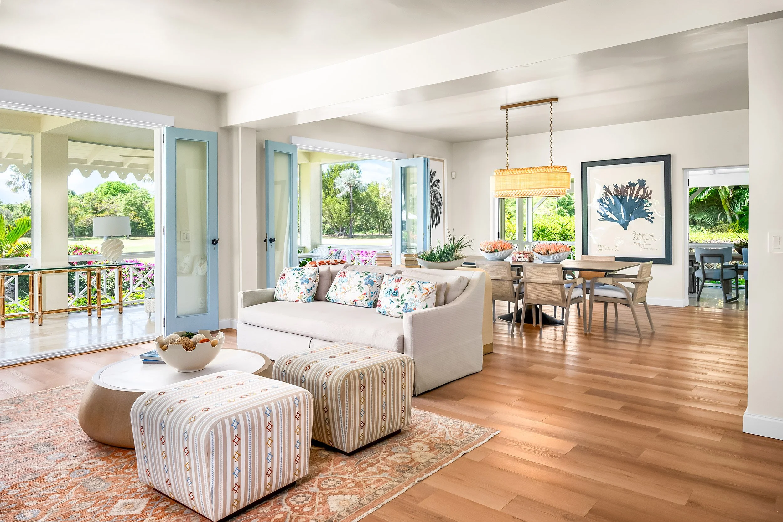 Living room with open doors leading to a balcony with a picnic table. Furniture includes a white sofa with patterned cushions, striped upholstered chairs, a round coffee table with a decorative bowl, and a dining table with chairs. Decor features framed botanical prints and floral arrangements. Large windows and glass doors provide views of lush greenery and palm trees outside. The floor is hardwood, and a textured rug is partially visible.