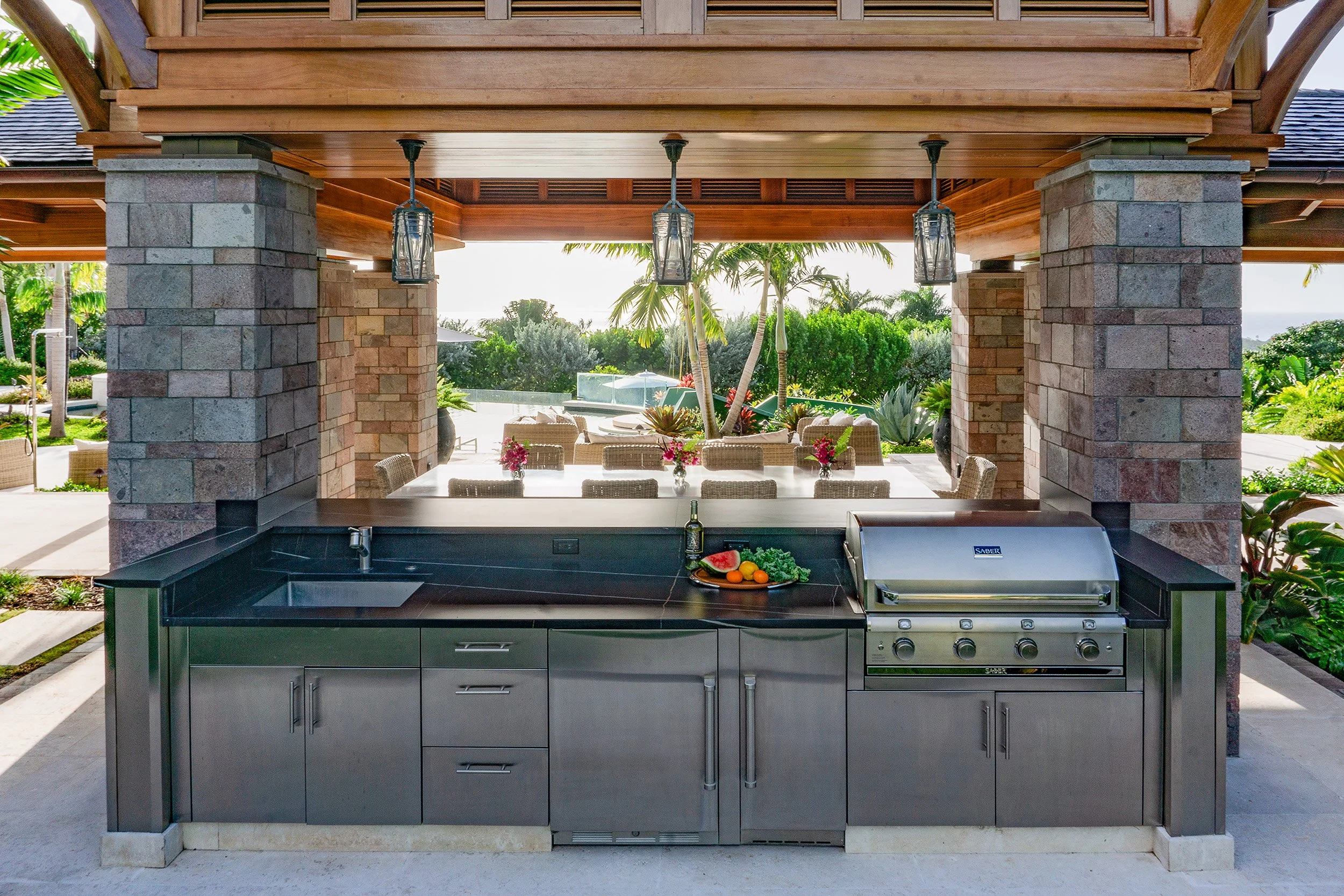 Outdoor kitchen with a black grill, countertop, and island with a sink, overlooking a lush garden with palm trees and tropical plants.
