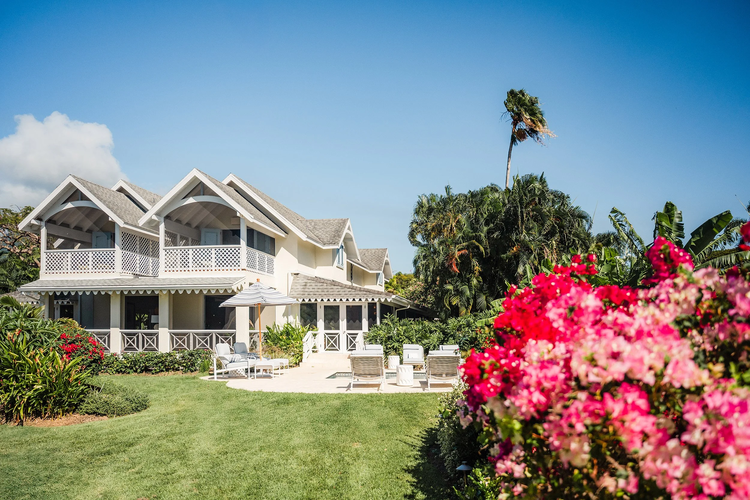 A white house with multiple gabled roofs, a porch, and outdoor seating, surrounded by lush greenery, pink flowers, and palm trees under a clear blue sky.