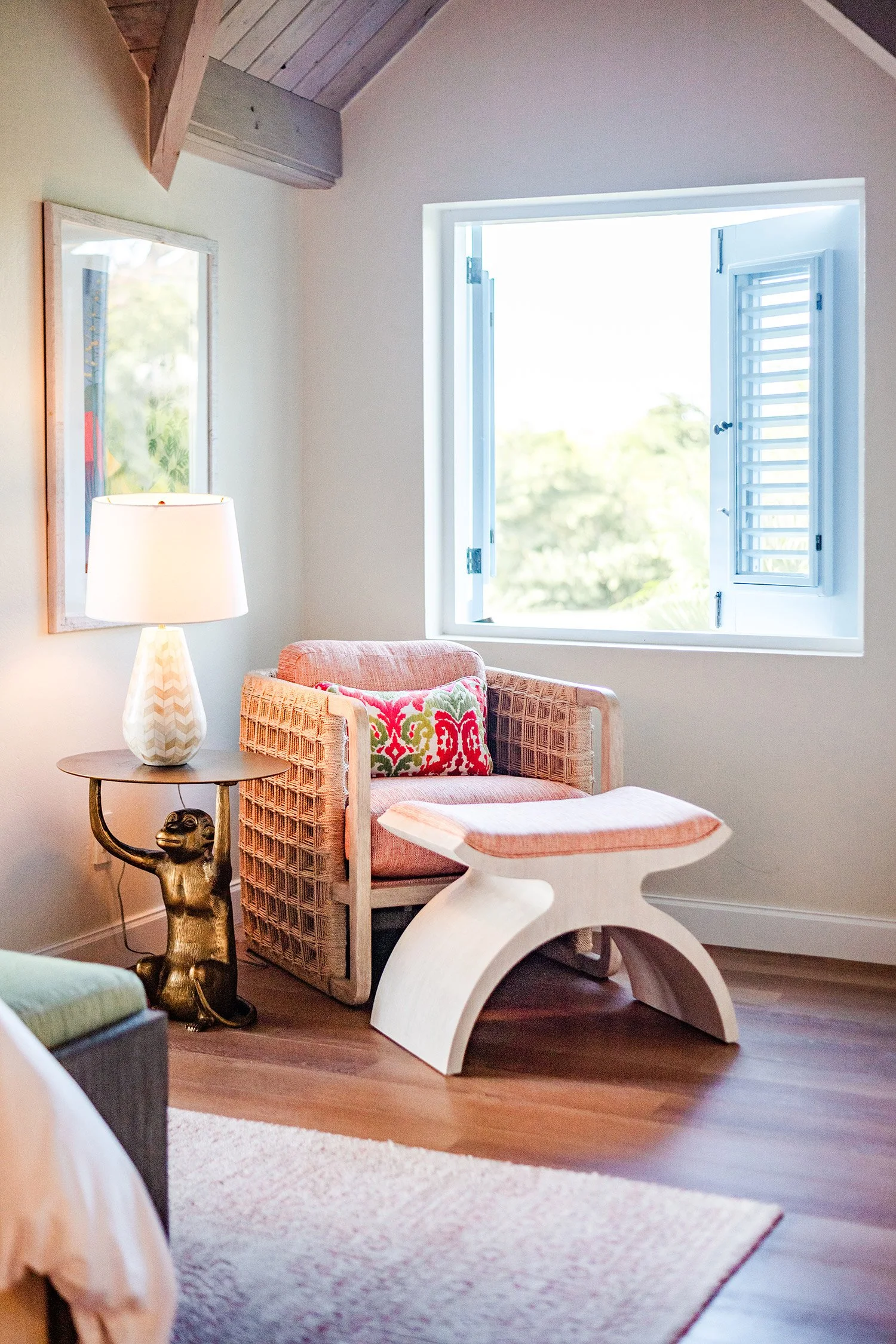 A cozy living room corner with a rattan armchair, a colorful cushion, a modern white side table, a white lamp with a patterned base, a decorative gold monkey figurine, an open window with shutters, a framed mirror, a wooden ceiling, and a light-colored rug.