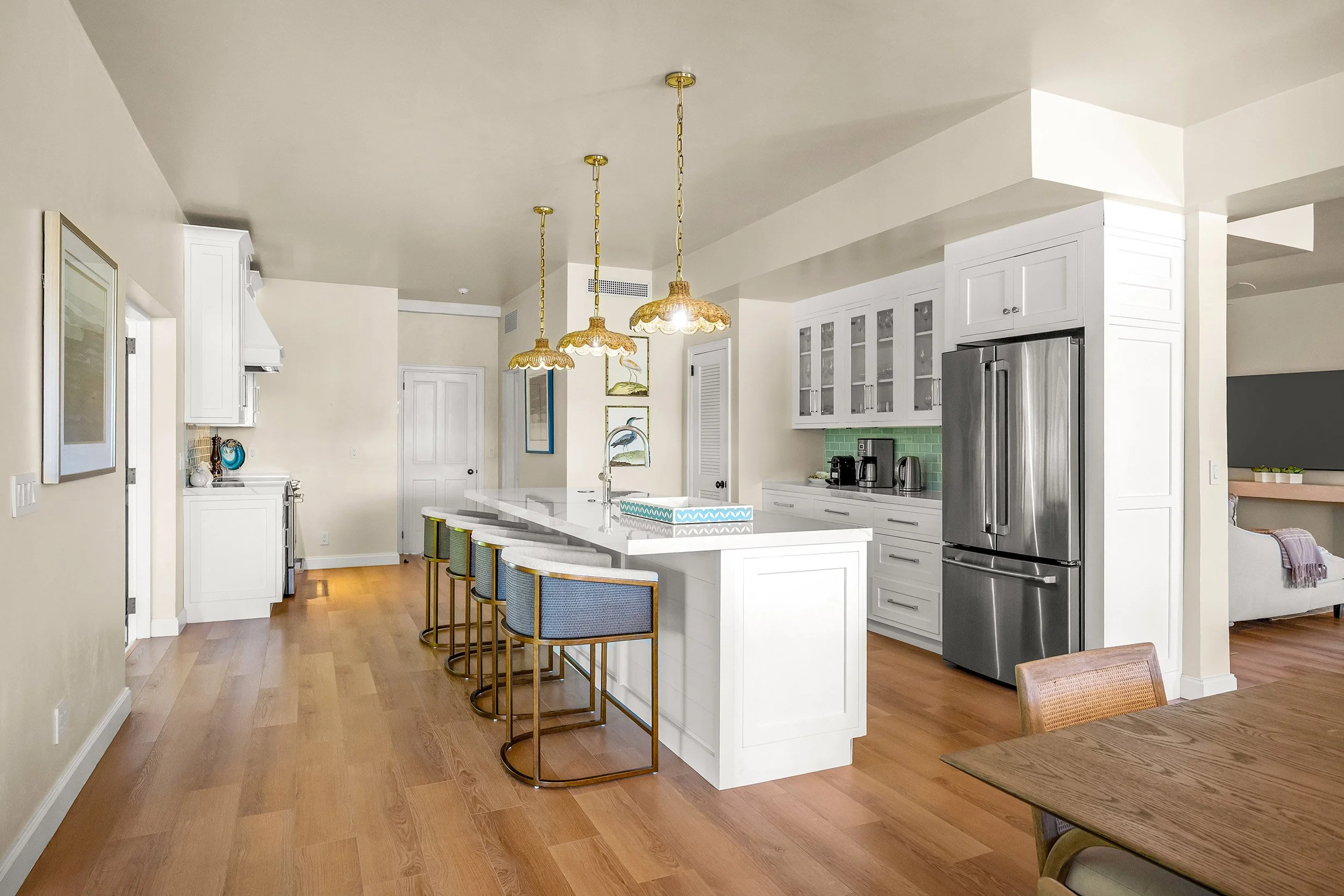 Modern kitchen with white cabinets, stainless steel refrigerator, green tiled backsplash, and a white island with four chairs, three hanging ceiling lights, and decorated with plants and artworks.