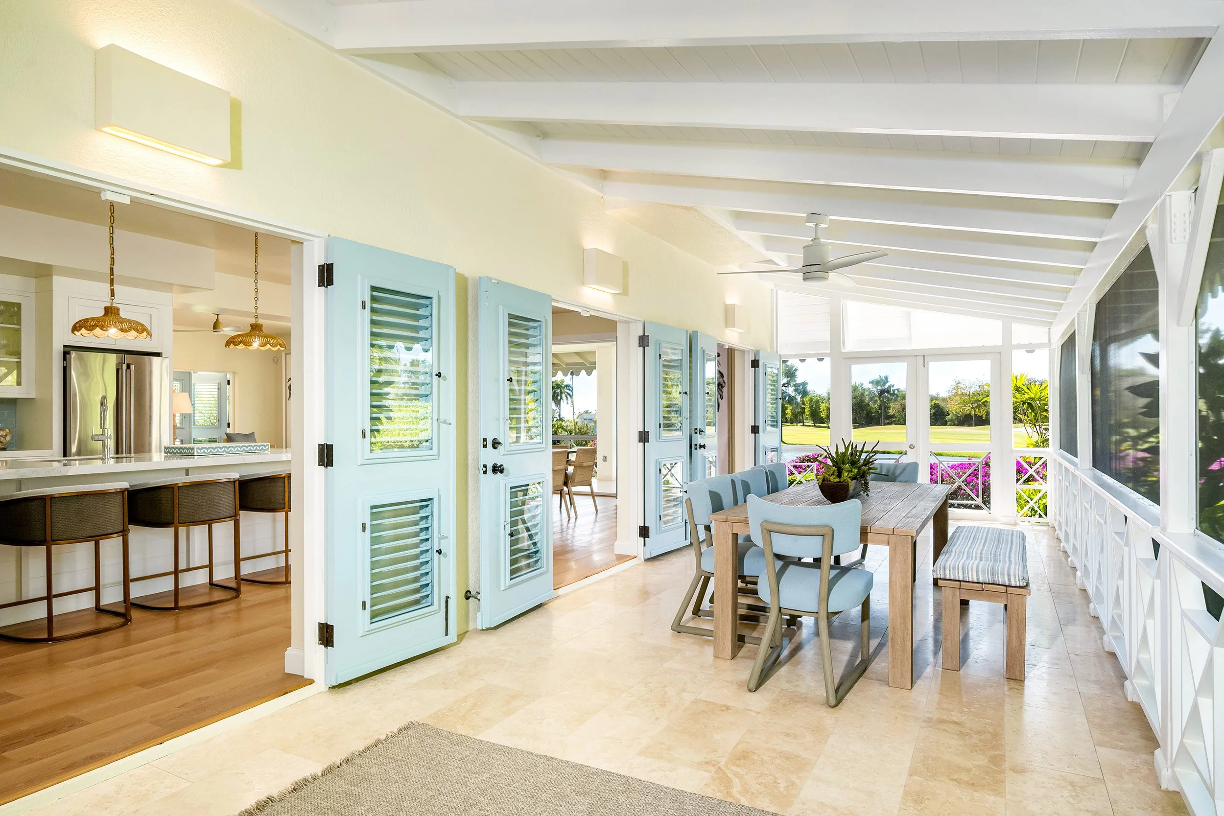 Bright sunlit enclosed porch with outdoor dining table and chairs, open shutters, and a view of greenery and a golf course outside.