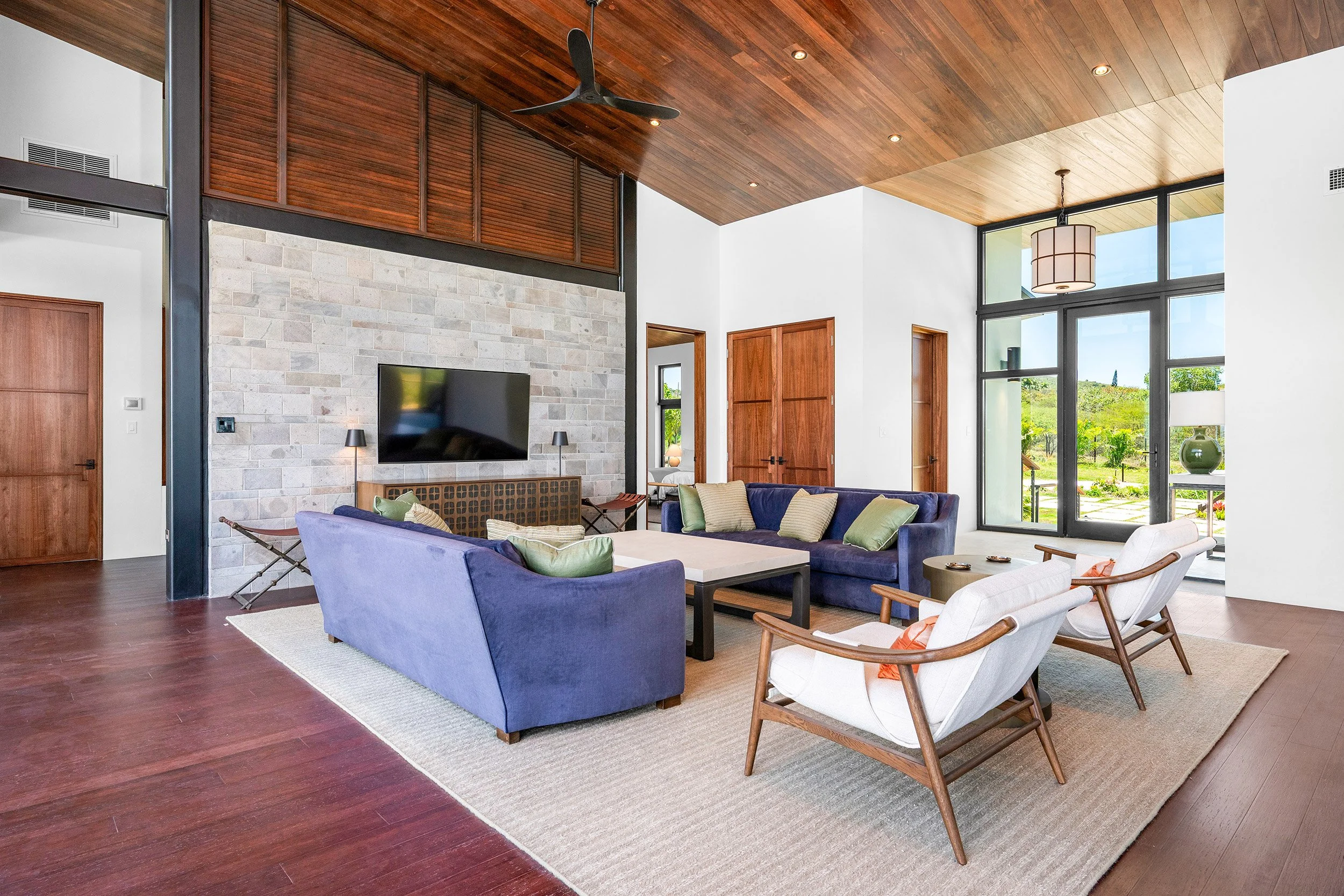 Modern living room with wooden ceiling, large glass windows, and white walls. It features a blue sofa with multiple cushions, a white armchair, a coffee table, and a tv mounted on a stone wall. There is also a wooden door, a ceiling fan, and a large hanging light fixture.