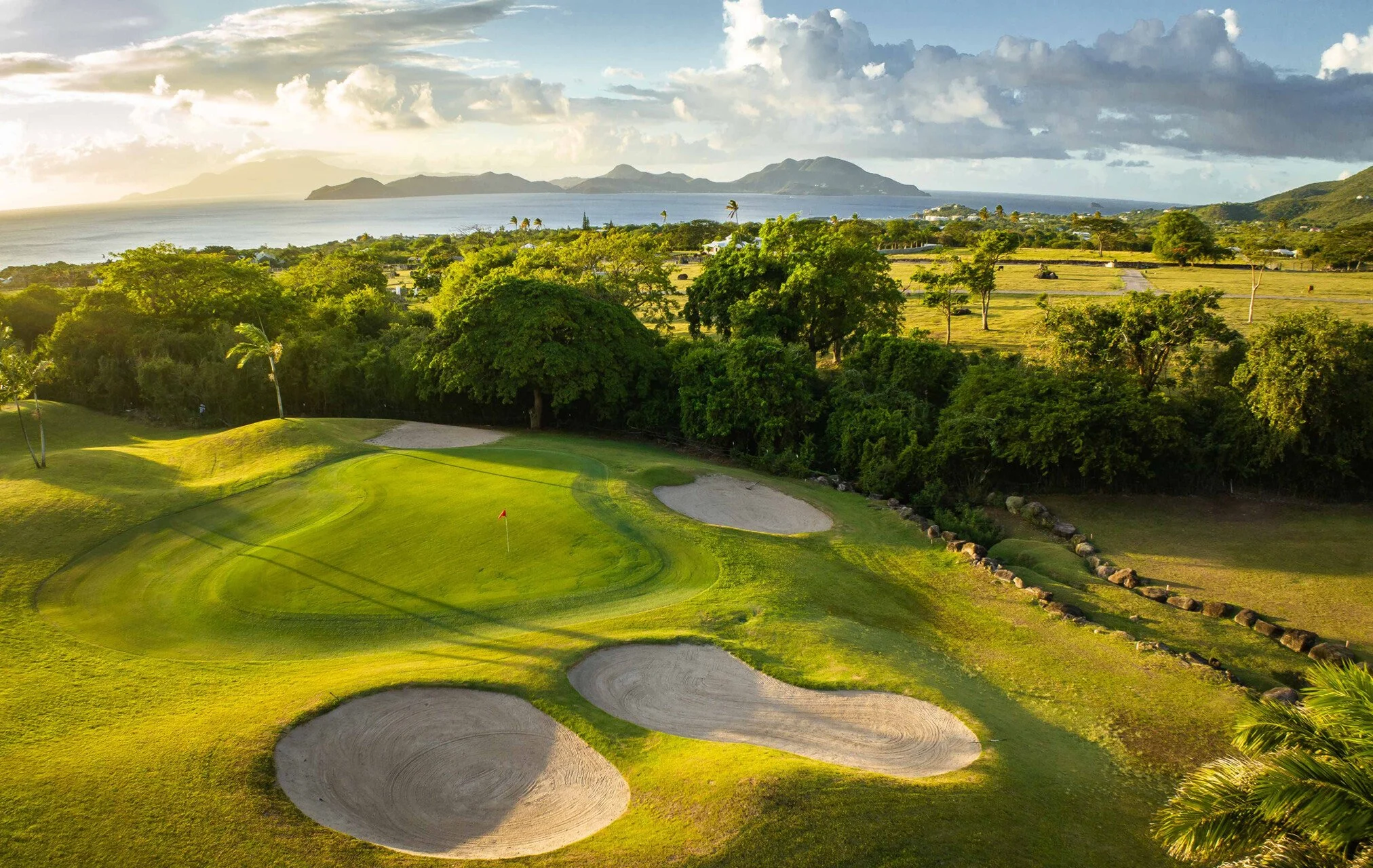 A golf course with well-maintained green fairways and bunkers, surrounded by trees and with an ocean and mountainous landscape in the background.