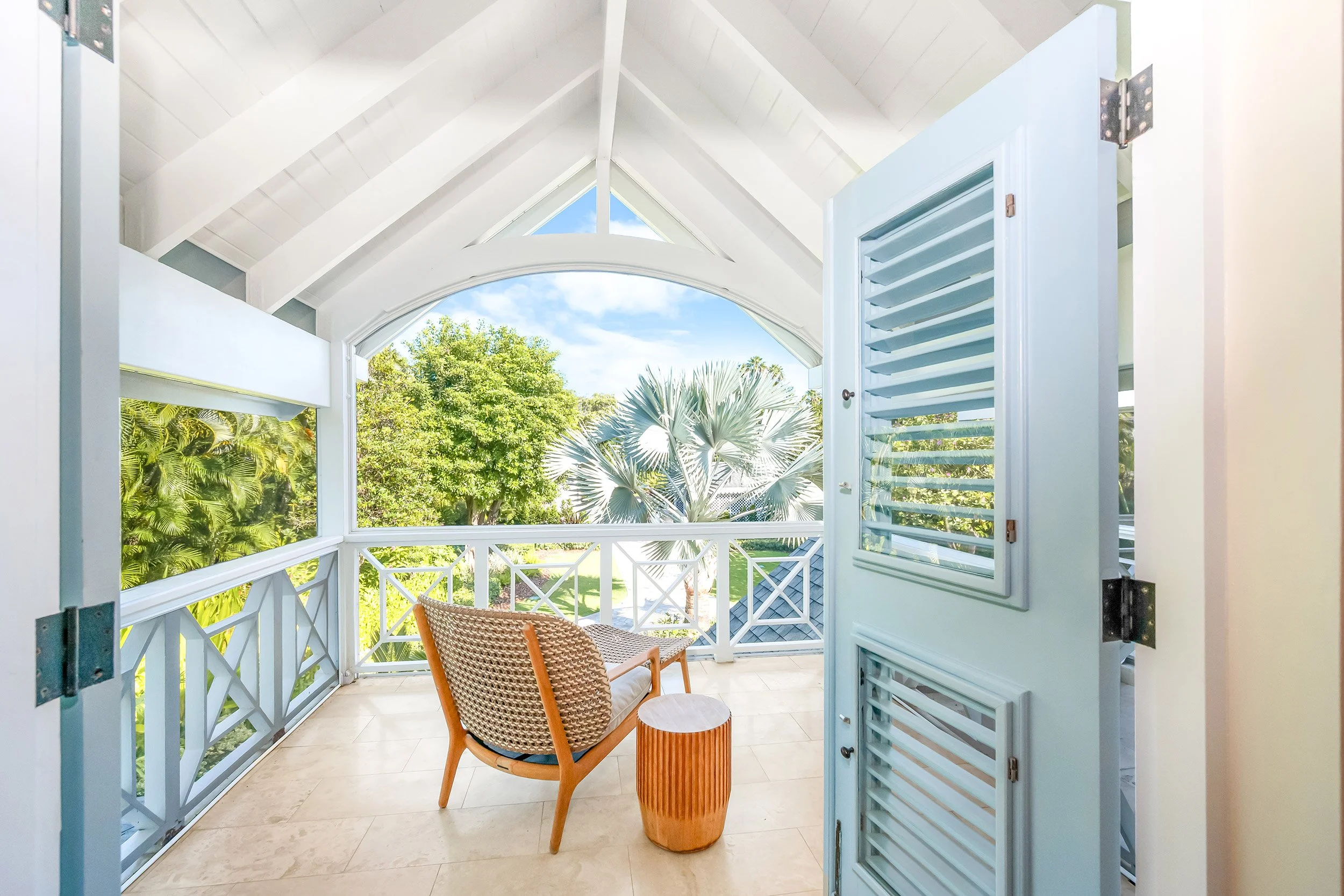 A porch with white framing, a wicker chair, and a small wooden side table, overlooking tropical trees through open shutters.