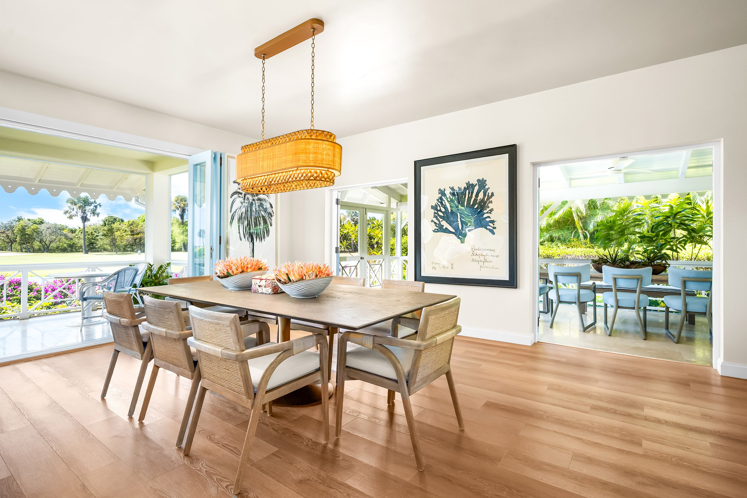 Bright living room with open doors leading to garden, wooden dining table, wicker light fixture, framed coral print, and views of greenery and trees outside.