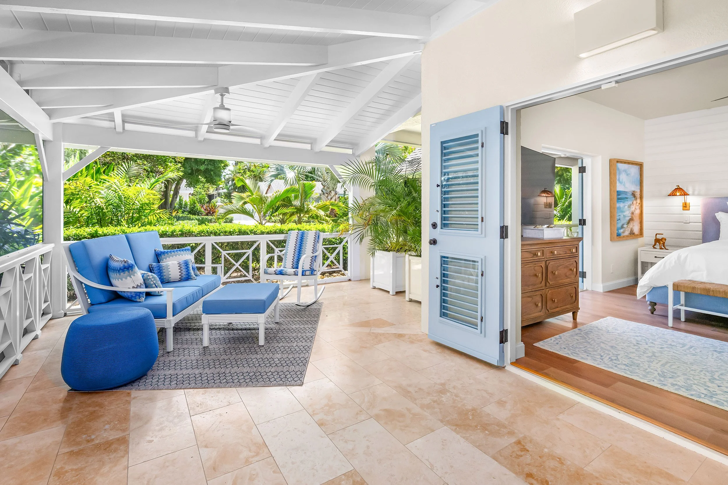 Interior view of a bright porch area with white wooden ceiling beams, a ceiling fan, blue outdoor furniture with cushions, and lush green plants outside. Adjoining room with a bed, wooden dresser, and artwork on the wall.