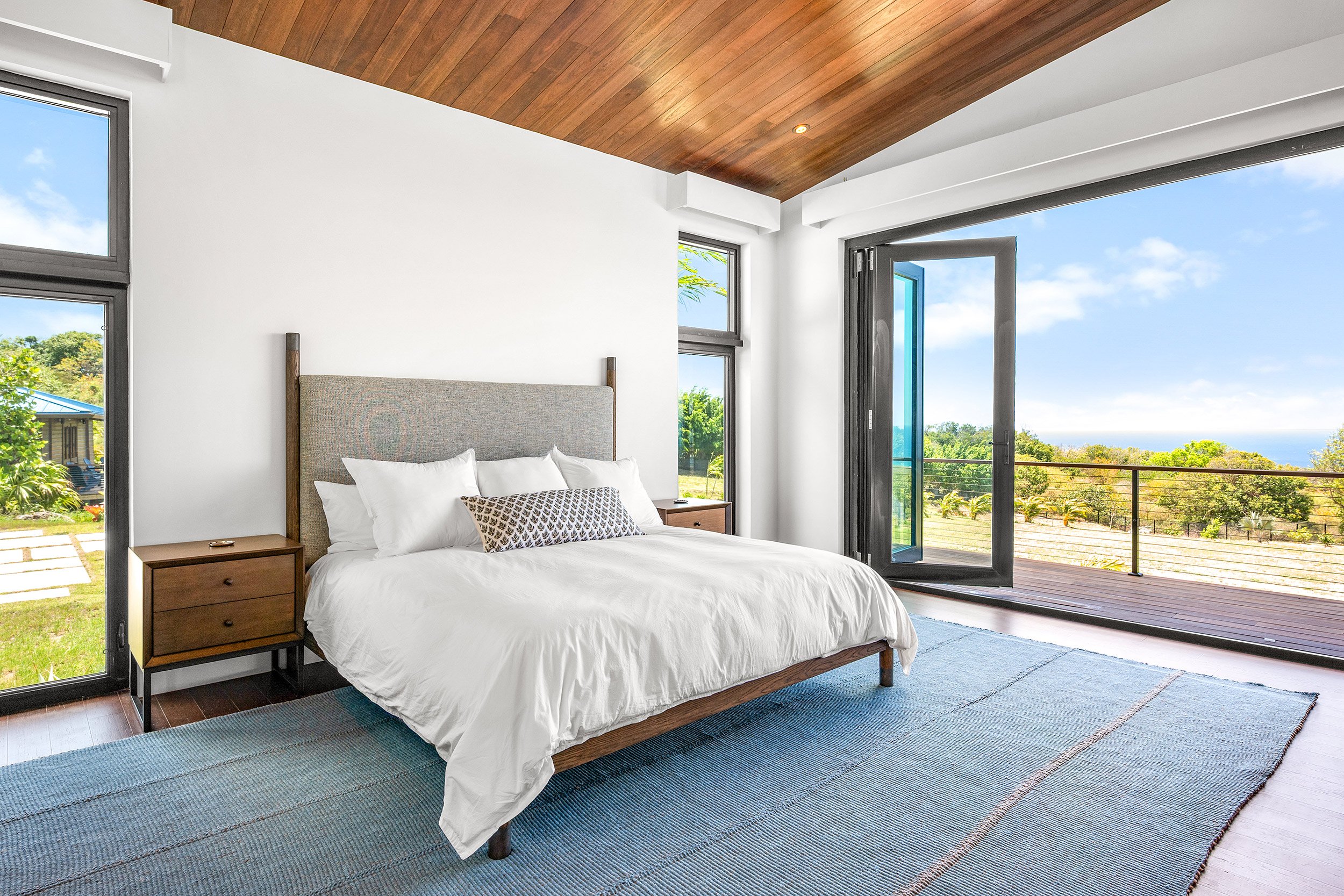 Modern bedroom with a large bed, wooden headboard, white bedding, and throw pillows. Large windows and a sliding glass door open to a balcony with a scenic view of trees and the ocean. Wooden ceiling and a blue striped rug on the floor.