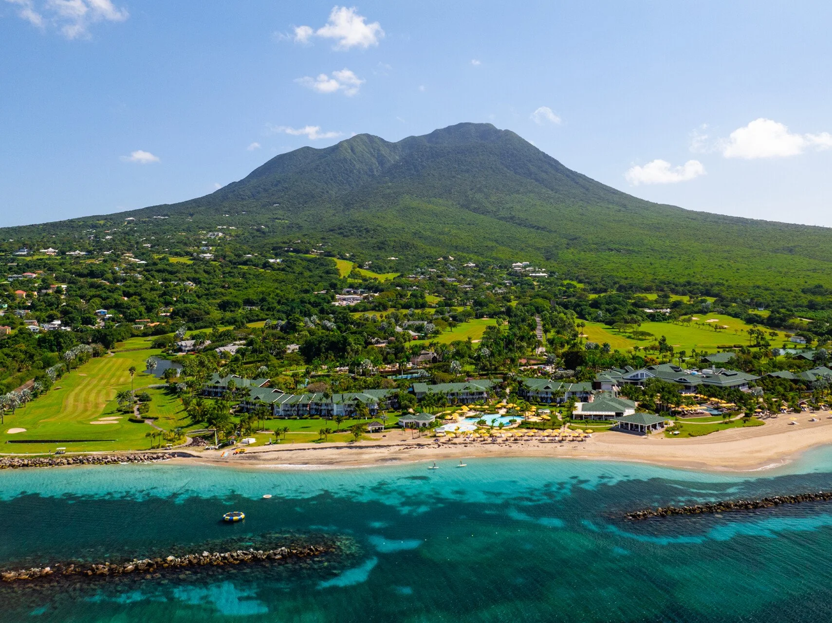 Aerial view of a tropical island with a mountain, green landscape, residential buildings, a golf course, a beach, and turquoise ocean water.
