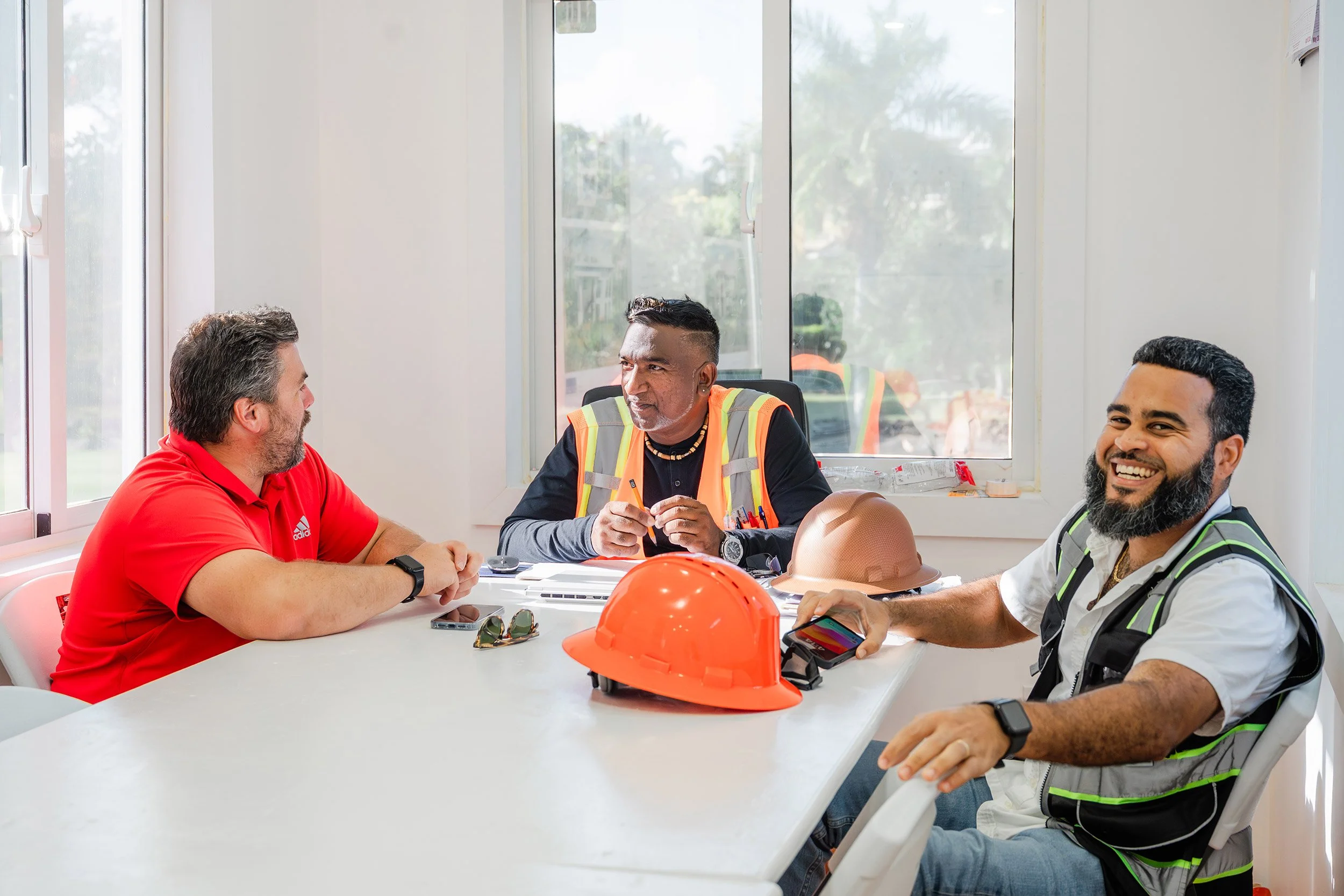 Three men sitting at a table engaged in conversation, with construction helmets and safety vests on the table, in front of a large window with a view of trees outside.