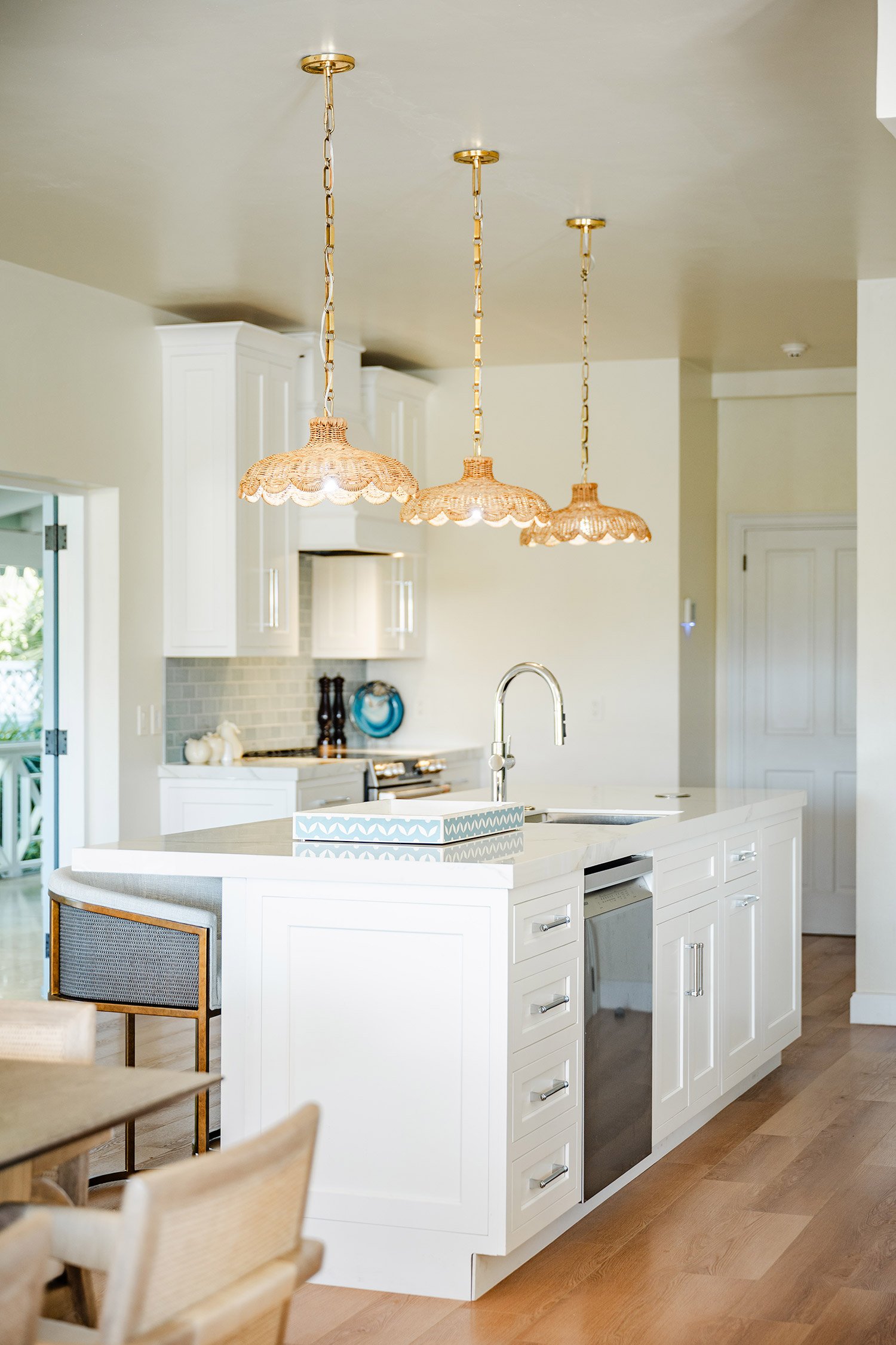 A modern kitchen with a white island featuring cabinet storage, a built-in microwave, and a sink. Above, there are three pendant lights with ruffled, wicker-like shades hanging from the ceiling. In the background, white kitchen cabinets, a stove, and decorative items on the counter are visible.