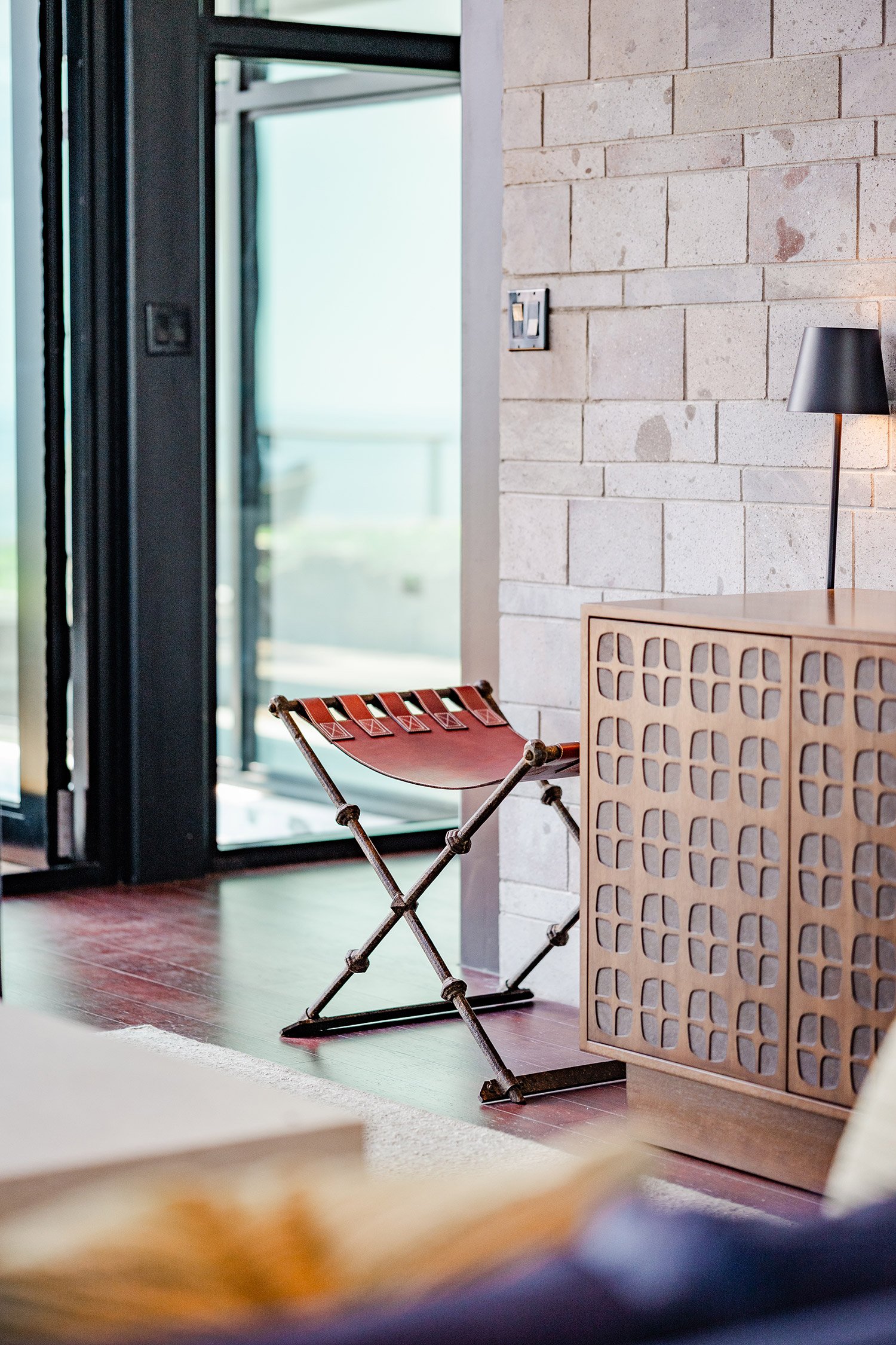Interior of a modern room with a brick wall, a black floor lamp, and a red foldable chair near a wooden cabinet with decorative cutouts. A sliding glass door leads outside.