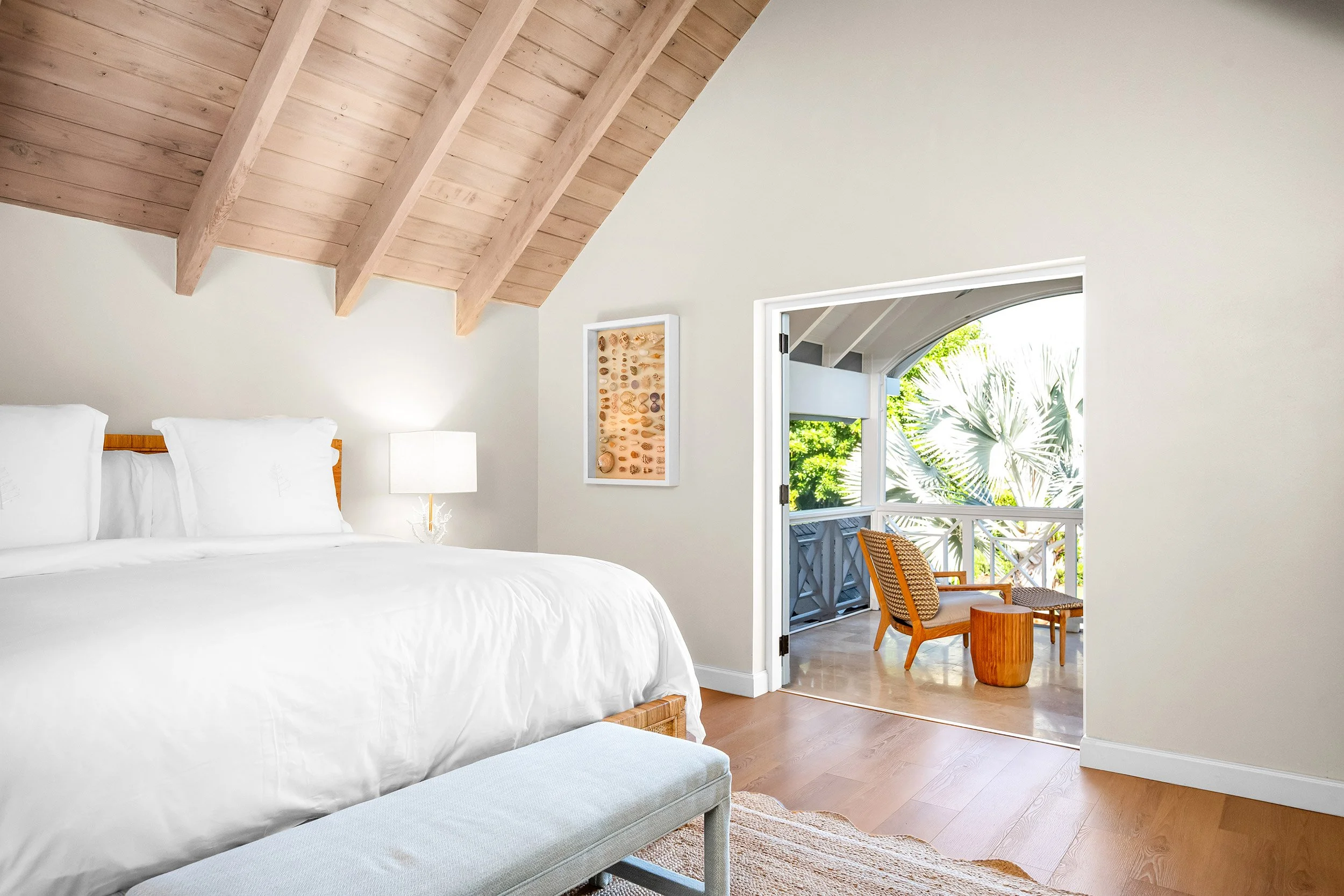 A bedroom with a white bed and a wooden bench at the foot of the bed. There's a nightstand with a white lamp and art with shells on the wall. A door opens to a balcony with outdoor seating and view of greenery.