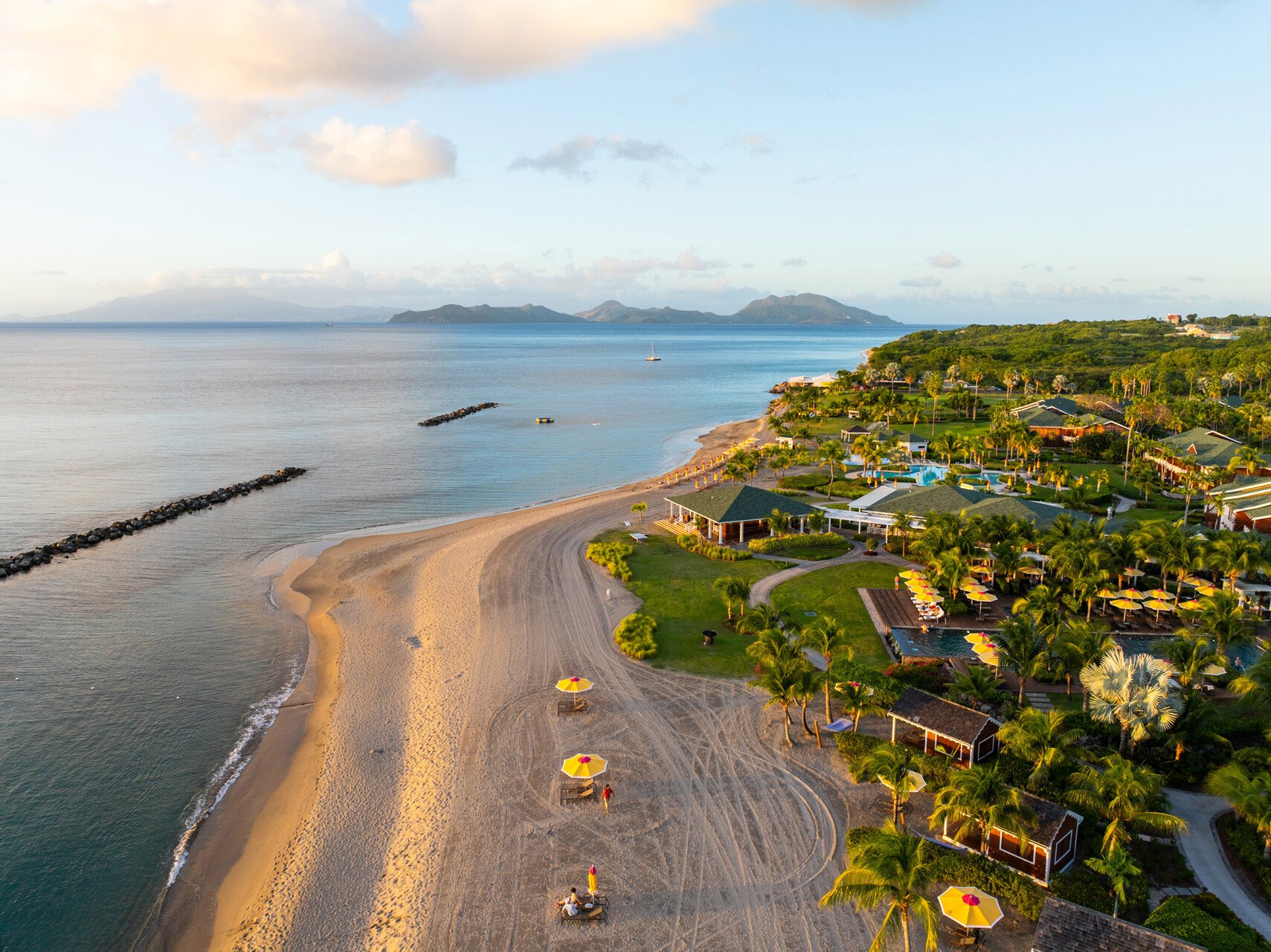Aerial view of a tropical beach resort with a sandy shoreline, palm trees, several buildings, swim pools, and yellow umbrellas along the beach. Calm ocean water extends to distant mountains on the horizon, with a sailboat and a small boat in the water. The sky is partly cloudy and sunlit.