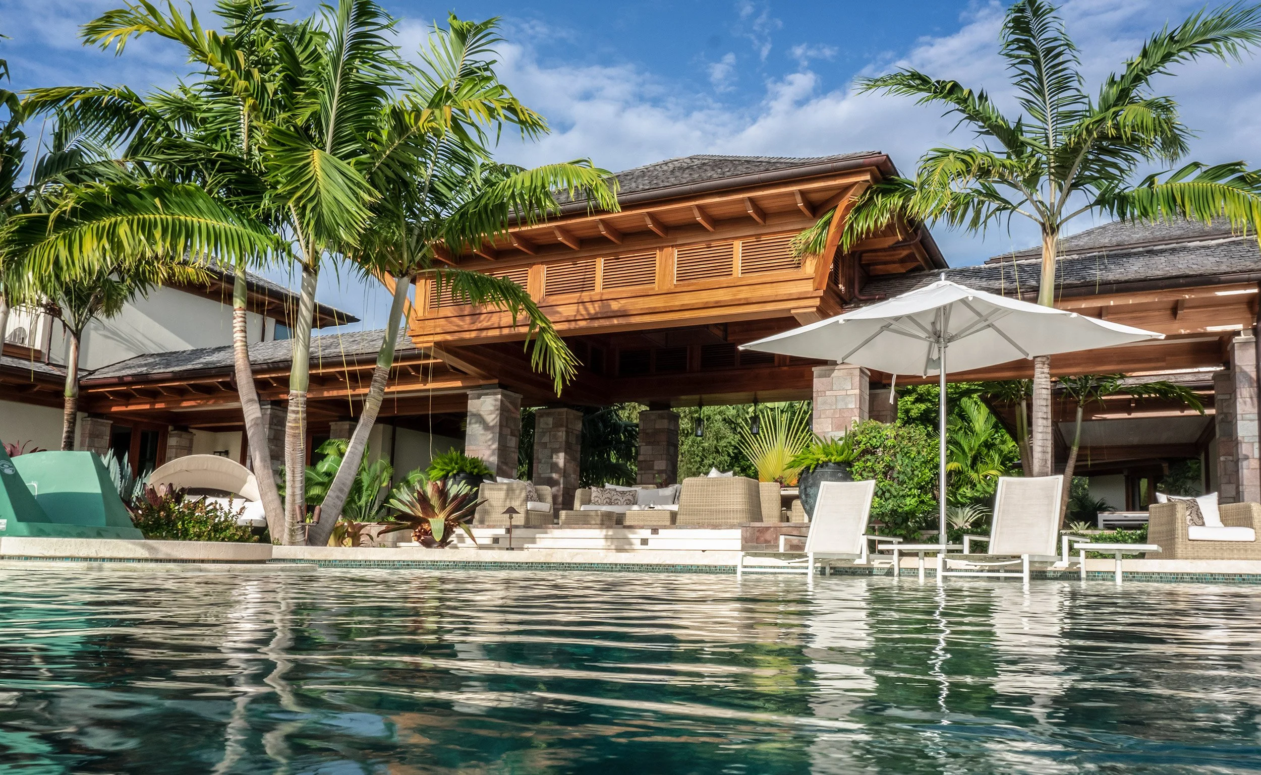A luxurious outdoor patio area beside a swimming pool, surrounded by palm trees and tropical plants, featuring wicker chairs, a white umbrella, and a wooden house with a covered deck under a partly cloudy sky.