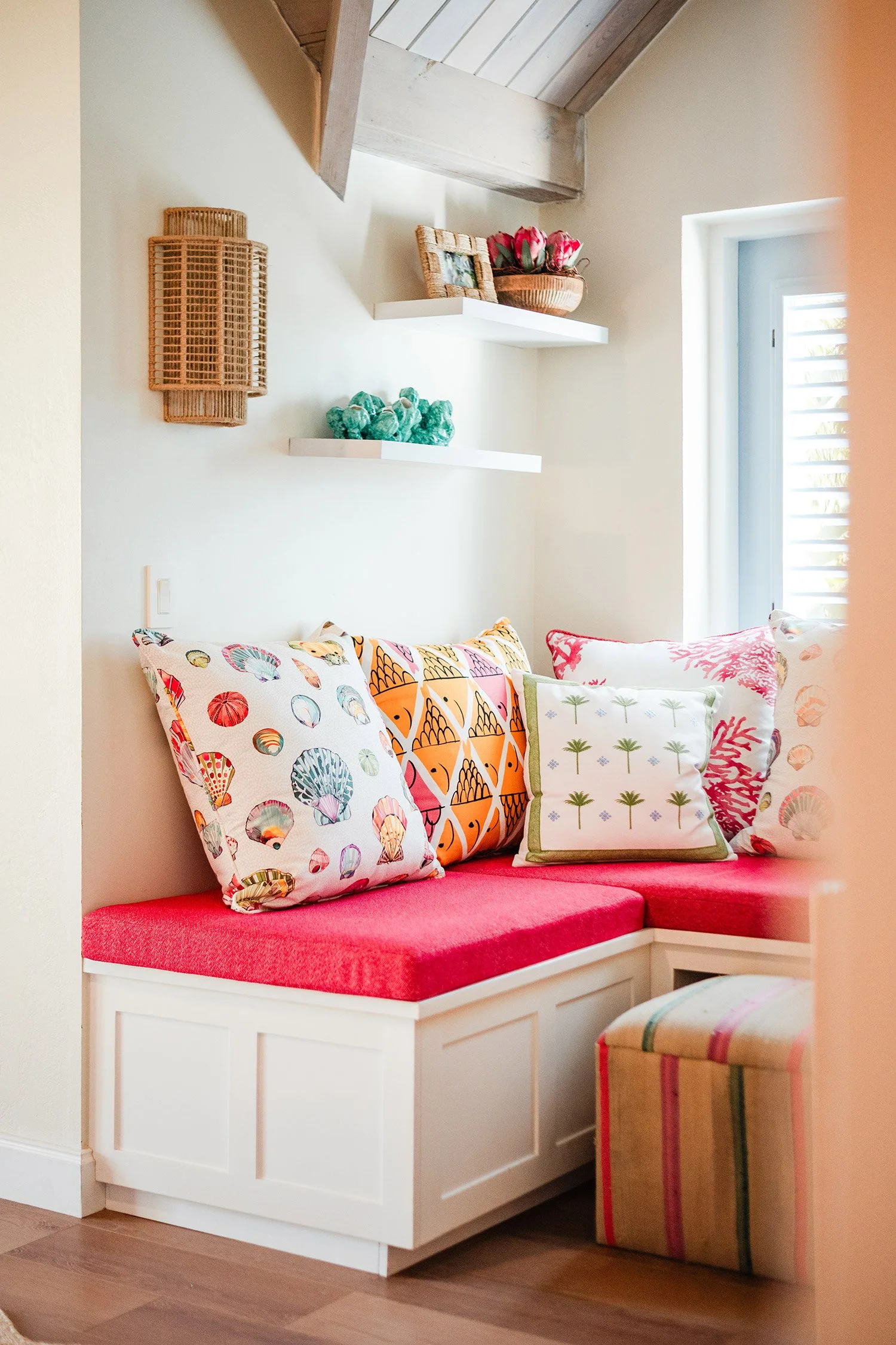A cozy corner with a built-in white bench featuring a red cushion, decorated with colorful throw pillows with various patterns including sea shells, fishes, and trees. There are floating white shelves above the bench holding a bowl of pink tulips, a small framed photo, and decorative green stones. Natural light filters in through a nearby window.