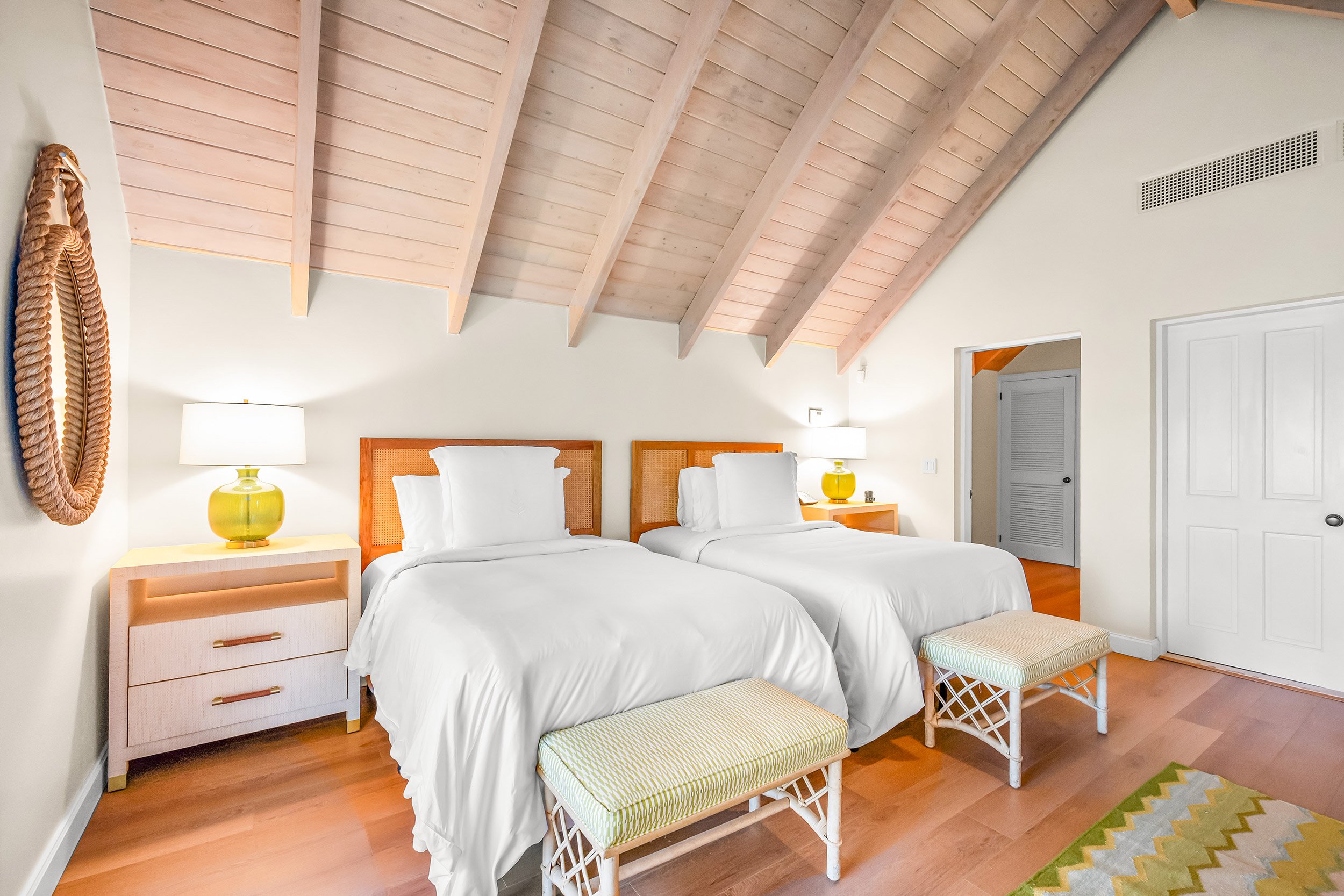 A cozy bedroom with two white beds with rattan headboards, side tables with yellow lamps, and a wooden ceiling with angled beams.