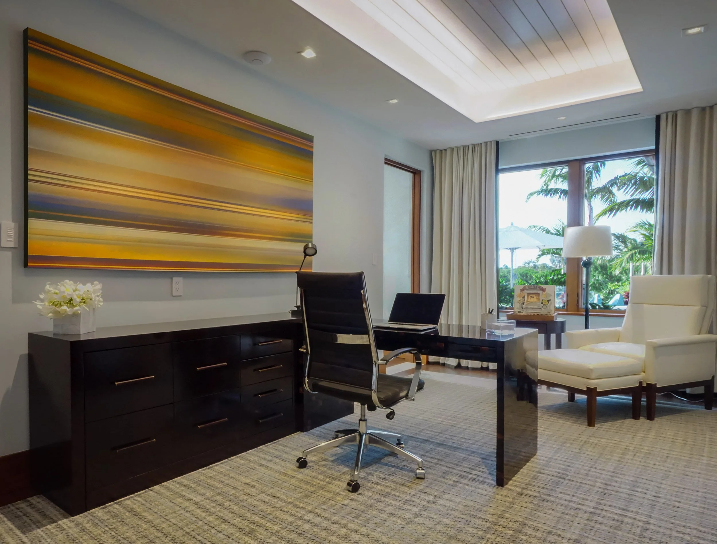 A modern office with a large window showing palm trees outside. The room contains a dark wooden desk, a black office chair, a white armchair, a tall floor lamp, and an abstract painting with horizontal yellow, orange, and purple streaks on the wall. There are cream-colored curtains on the window and a patterned carpet on the floor.