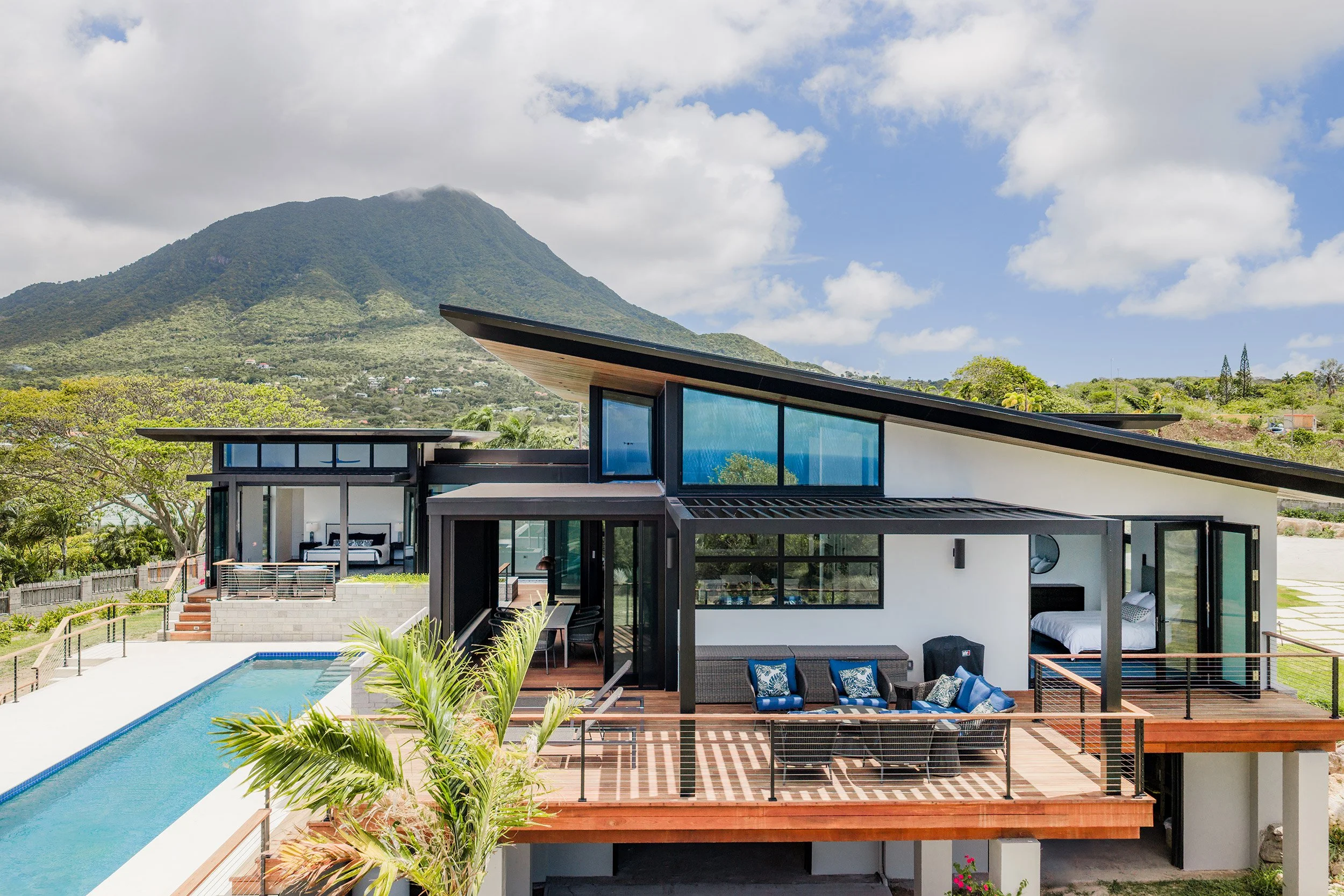 Modern house with flat roof, large glass windows, and an outdoor swimming pool, surrounded by lush greenery and with mountains in the background under a partly cloudy sky.