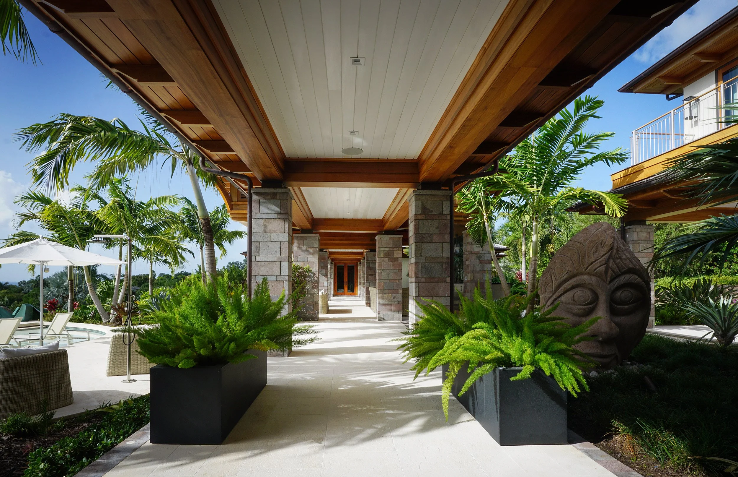 View of an outdoor patio area with a wooden ceiling, lush green plants, stone columns, and tropical landscaping in a backyard setting.