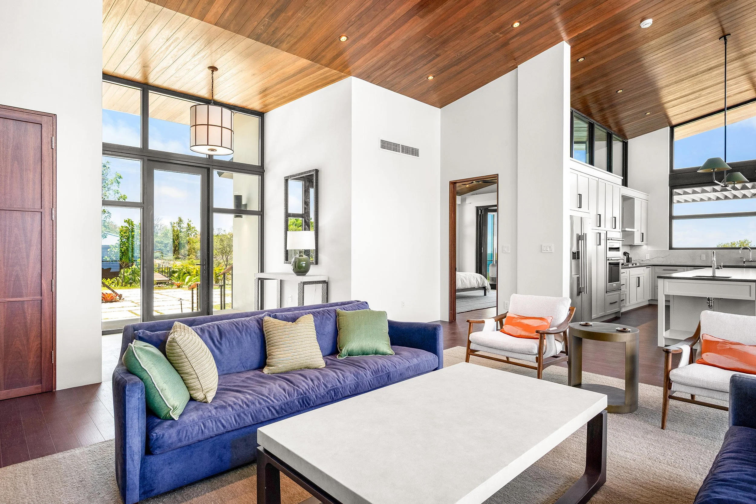 Living room with glass doors, white walls, wooden ceiling, and white kitchen cabinets, featuring a purple sofa with green and beige pillows, a white coffee table, and modern chairs.