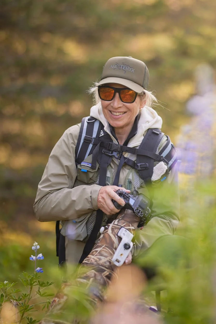 A woman in outdoor gear, wearing sunglasses and a cap, is smiling while holding a camera during a nature outing. She has a backpack with a tripod and a camouflaged spotting scope.