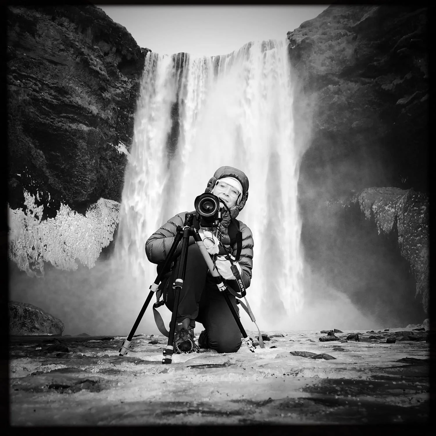 A person kneeling on a rocky surface in front of a waterfall, taking a photograph with a camera mounted on a tripod, wearing outdoor gear and glasses in black and white