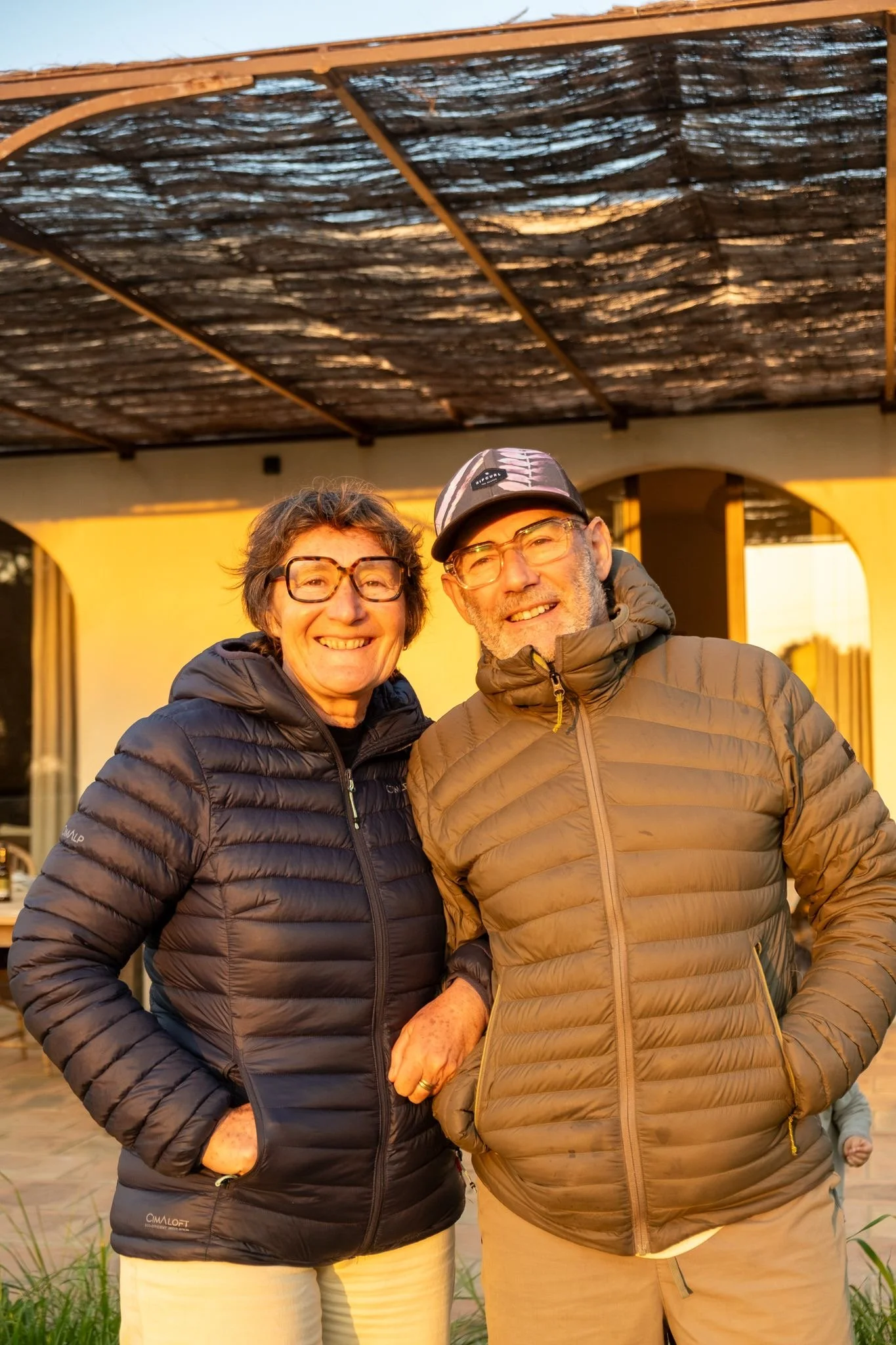 Un homme et une femme souriants portant des vestes matelassées, main dans la poche. Le coucher de soleil illumine leur visage, derrière une maison avec un porche.