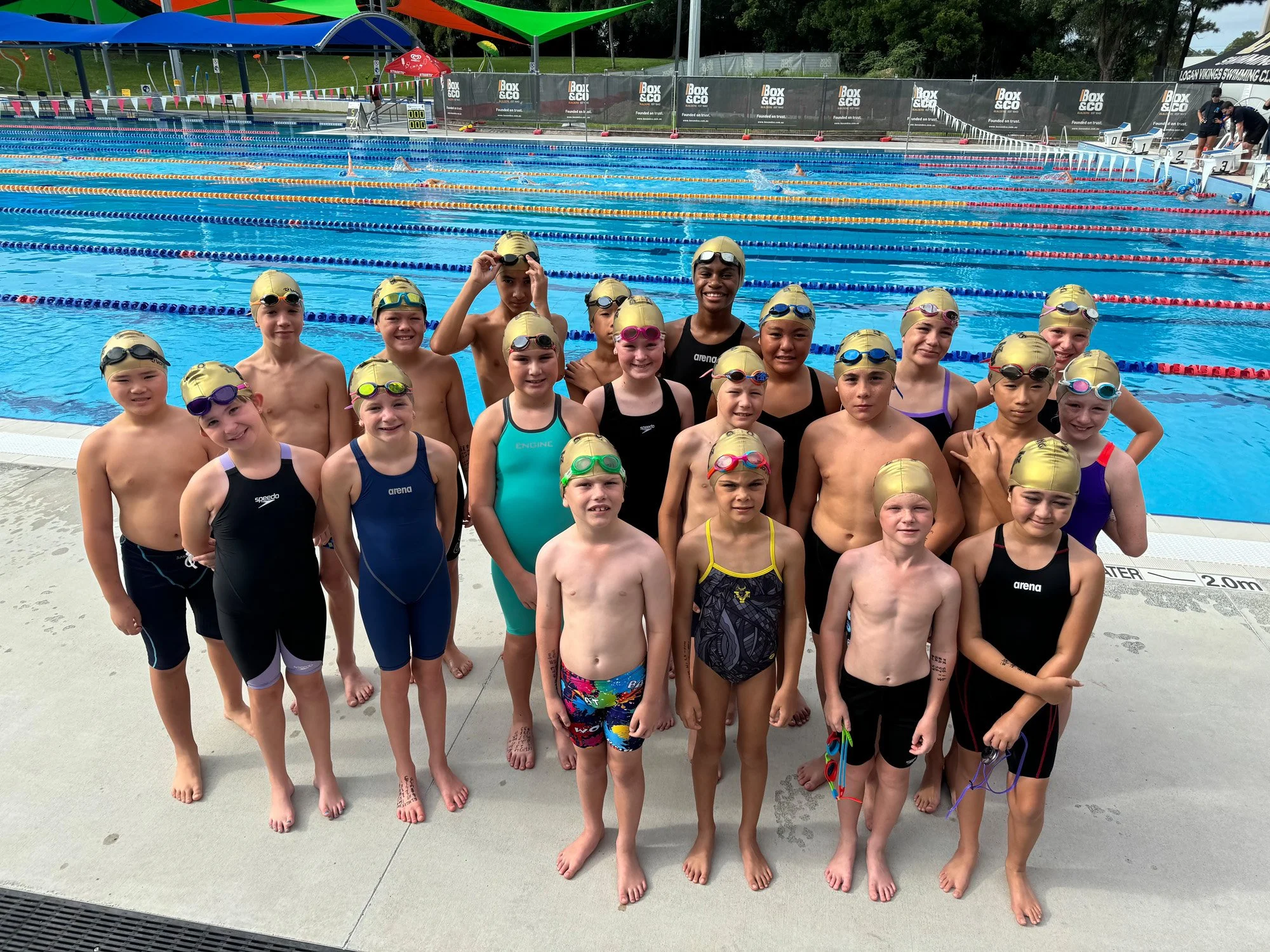 A group of young children and a coach gathers on the pool deck for a swim class photo at an outdoor swimming pool. They are wearing swim caps and goggles, and some are in swimsuits while others are in swim jammers or thin suits. The pool has multiple lanes separated by colorful lane markers, and there are some people swimming in the background.