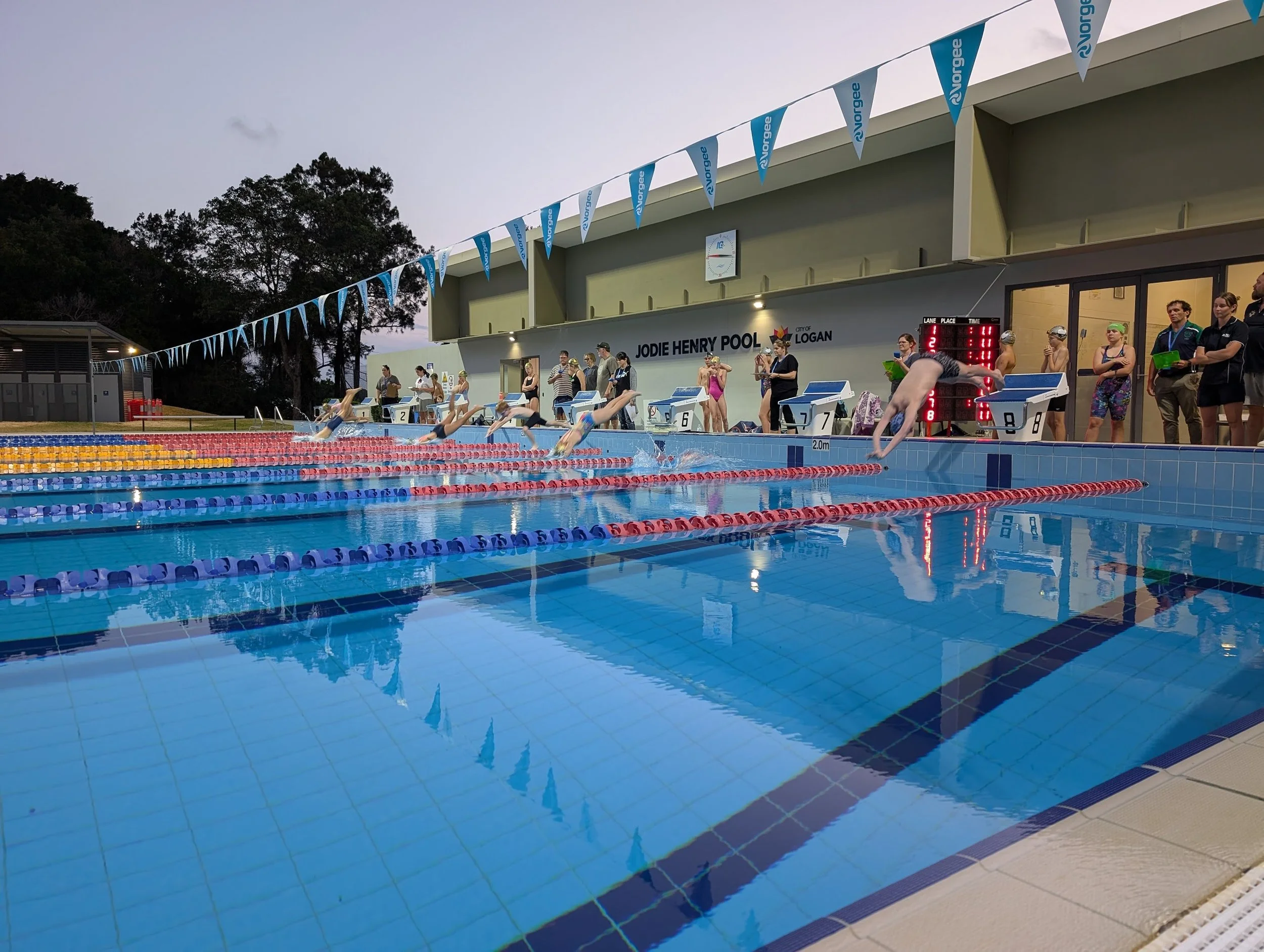 Swimmers diving into a pool during a race at Jodie Henry Pool with spectators watching from the sidelines.
