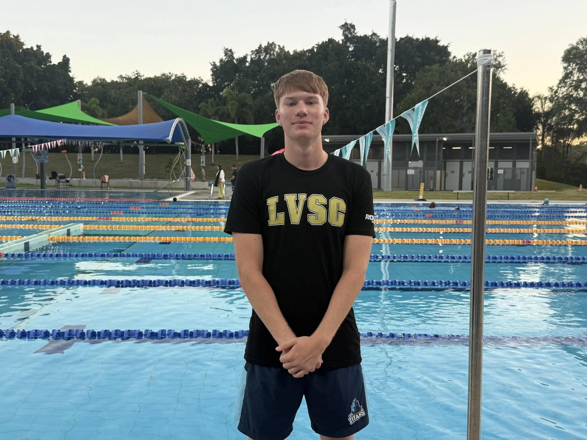 Young man standing poolside at an outdoor swimming pool during evening or early morning, wearing a black Lakers navy and yellow T-shirt and blue swimming shorts, with swimming lanes, poolside shade structures, and trees in the background.